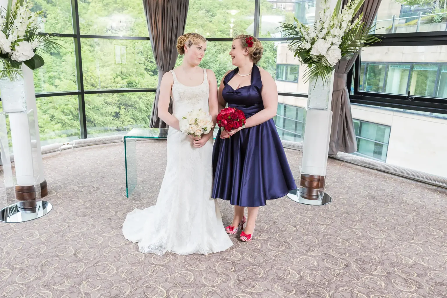 Two women at a wedding ceremony, one in a white bridal gown and the other in a dark blue dress, holding bouquets and standing in a room with large windows and city view.