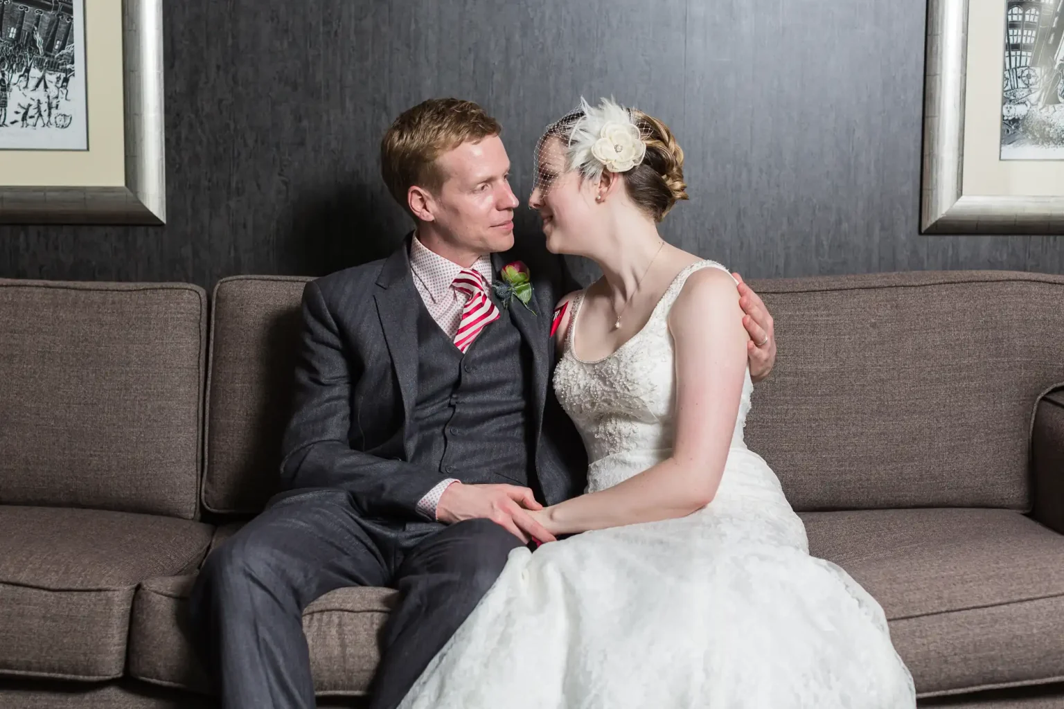 A newlywed couple sitting closely on a sofa, lovingly gazing at each other, the man in a suit and the woman in a white wedding dress.