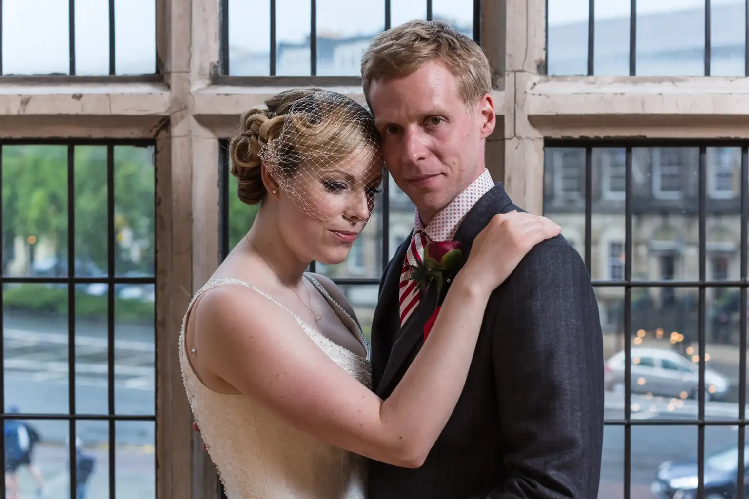 A bride and groom smiling and embracing each other by a window, with the bride wearing a veil and the groom in a gray suit and striped tie.