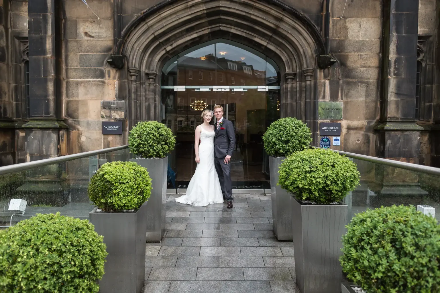 A bride and groom walking out of a church entrance, smiling and holding hands, with topiary plants lining the pathway.