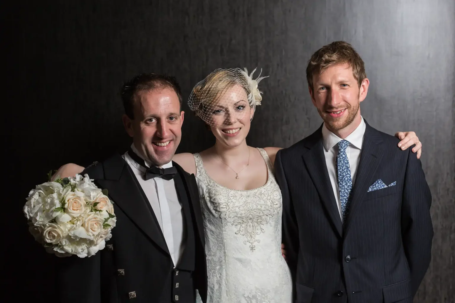 A bride in a white lace dress with a bouquet stands between two men in suits, one in a tuxedo, against a dark backdrop.