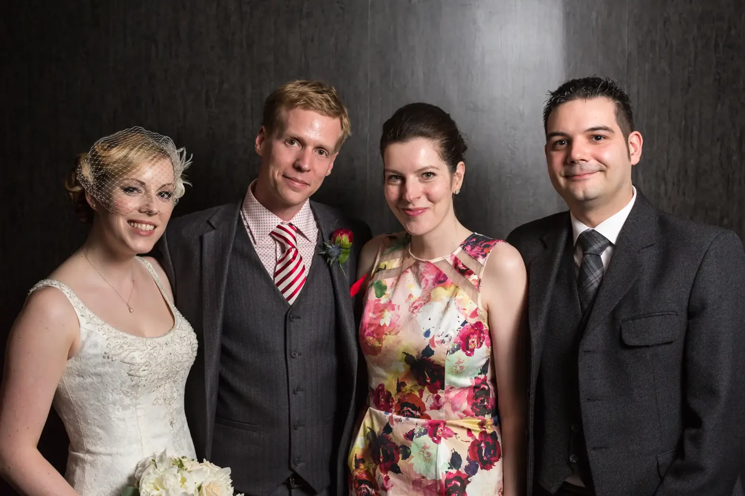 Two couples posing together, one in wedding attire and the other in formal wear, in front of a dark backdrop.