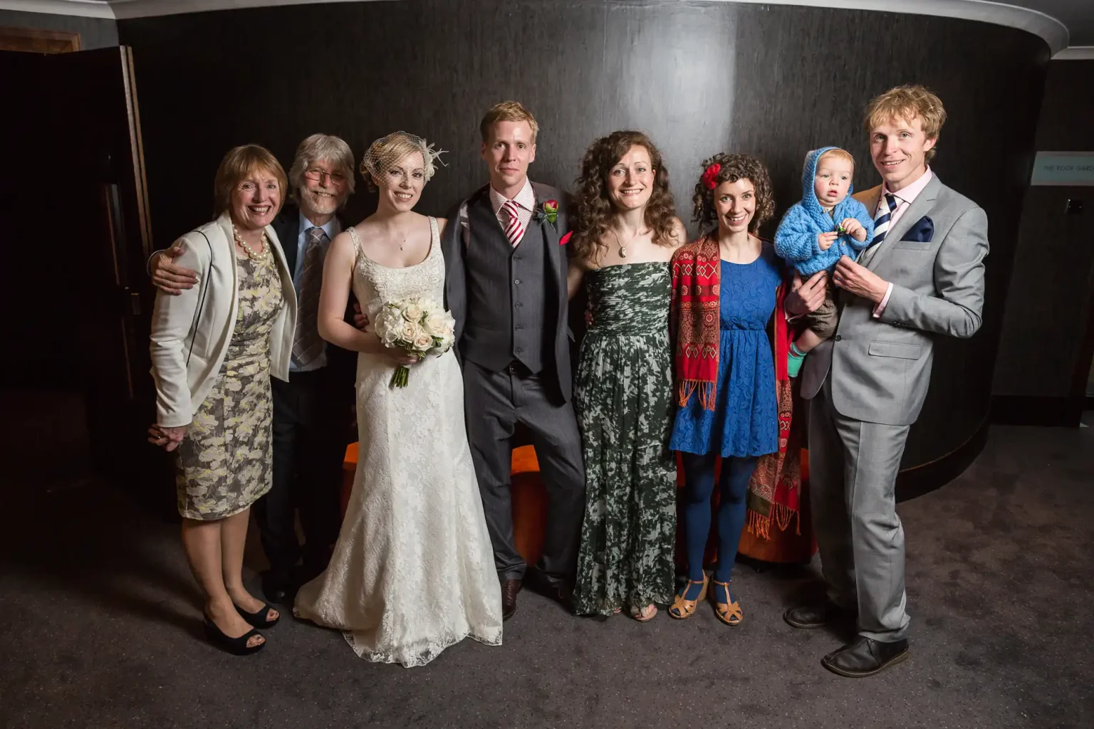A group of seven adults and one toddler posing at a wedding, smiling in formal and semi-formal attire.