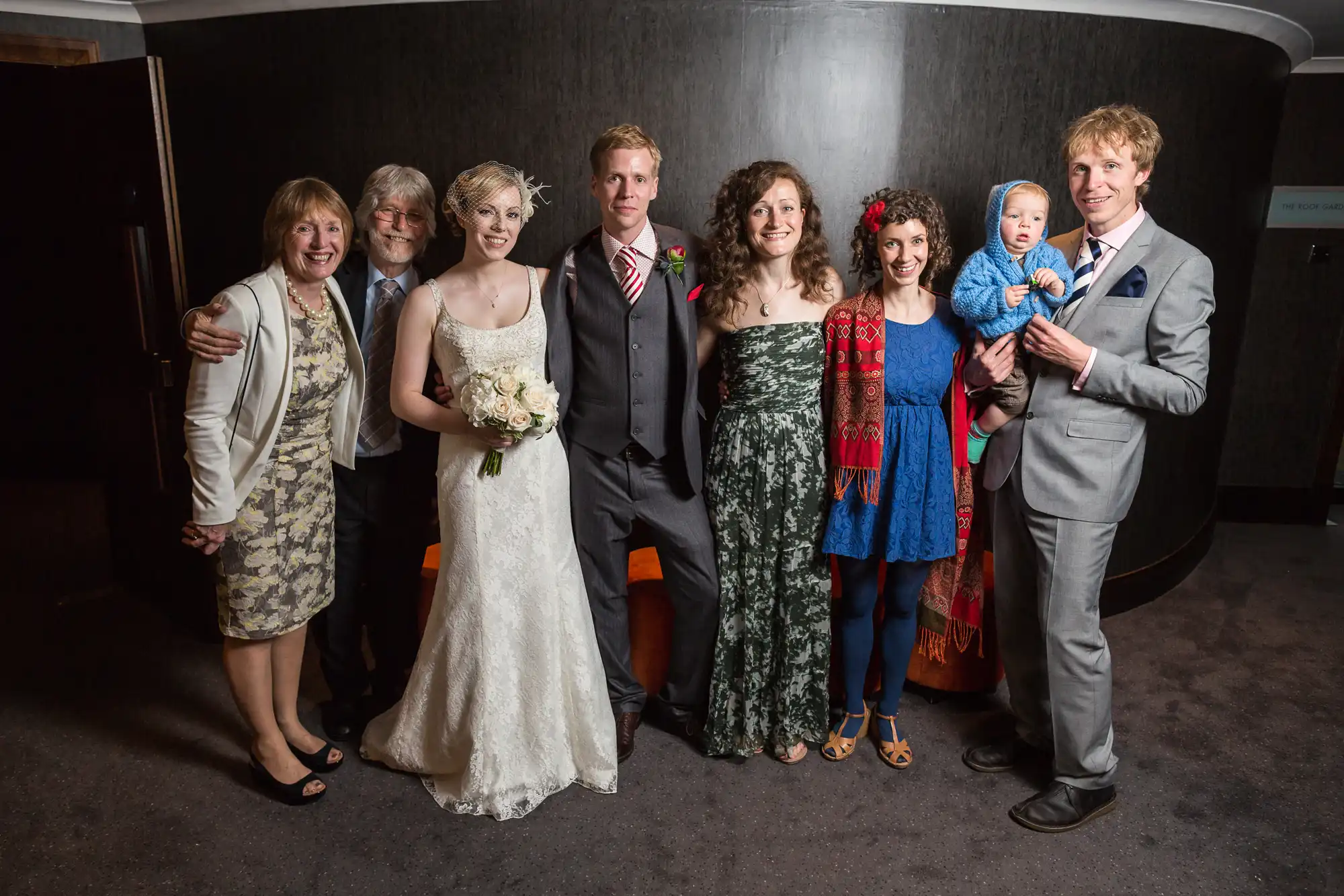 A group of seven adults and one toddler posing at a wedding, smiling in formal and semi-formal attire.
