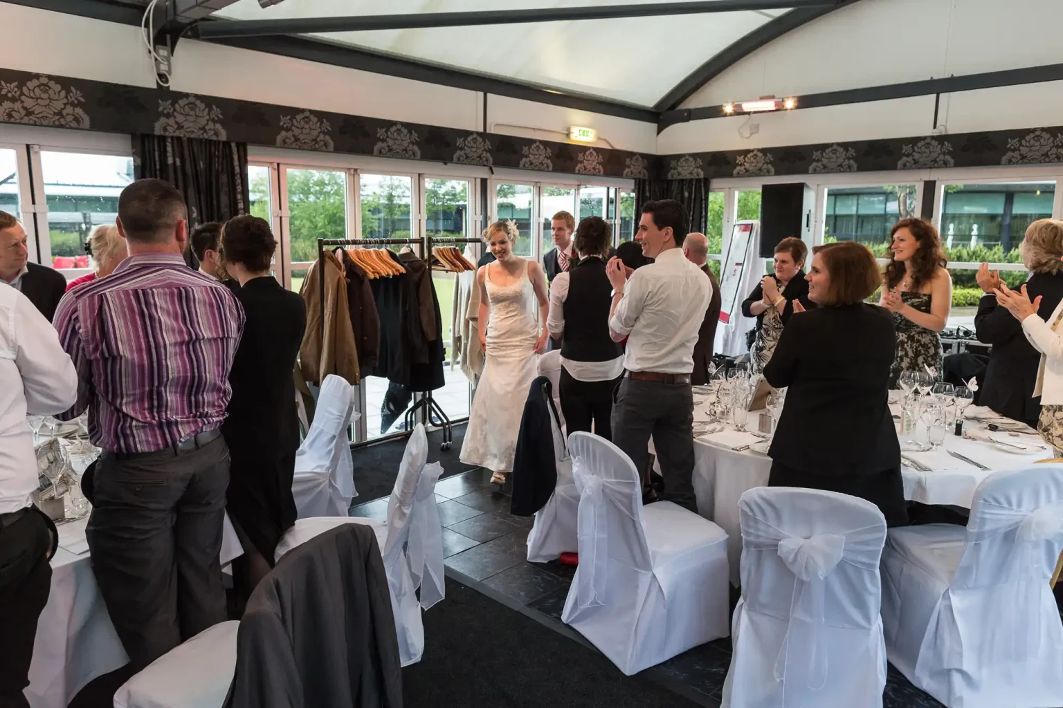 A bride and groom entering a wedding reception as guests applaud, decorated tables and chairs around them.