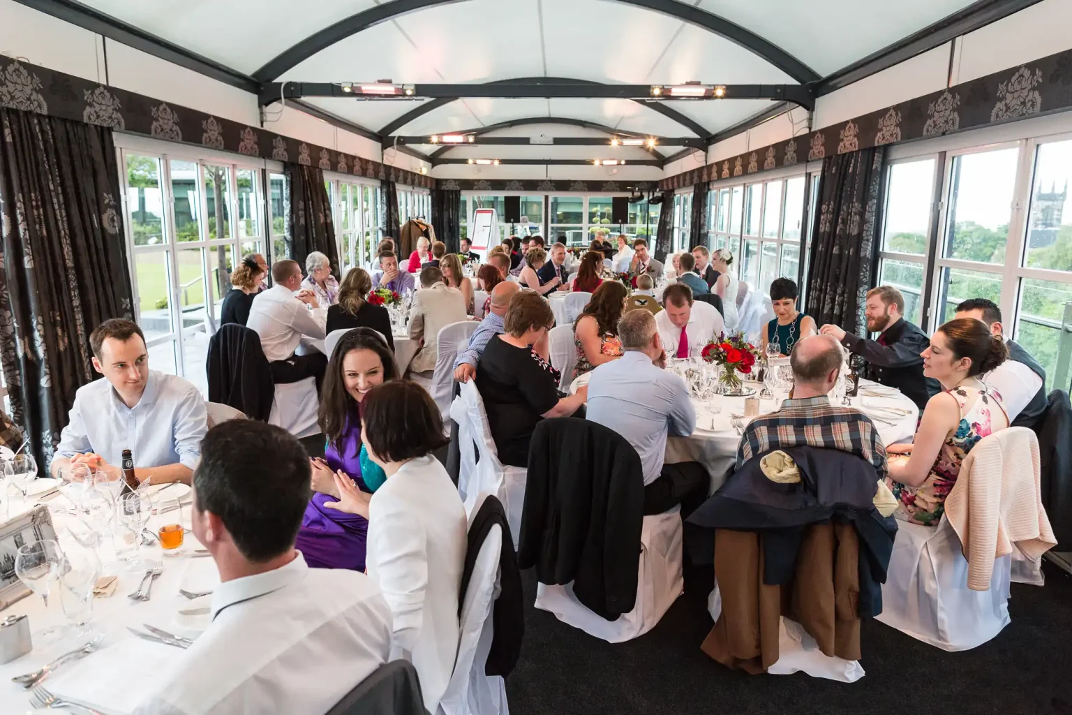 Diverse group of people dining in an elegant, elongated Calton Suite at The Glasshouse, with large windows and patterned decor.