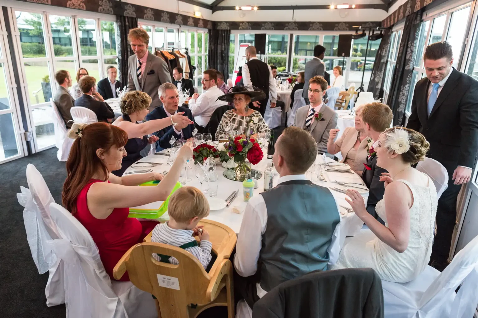 Guests seated at a wedding reception, engaged in conversation and dining, in an elegantly decorated venue with large windows.