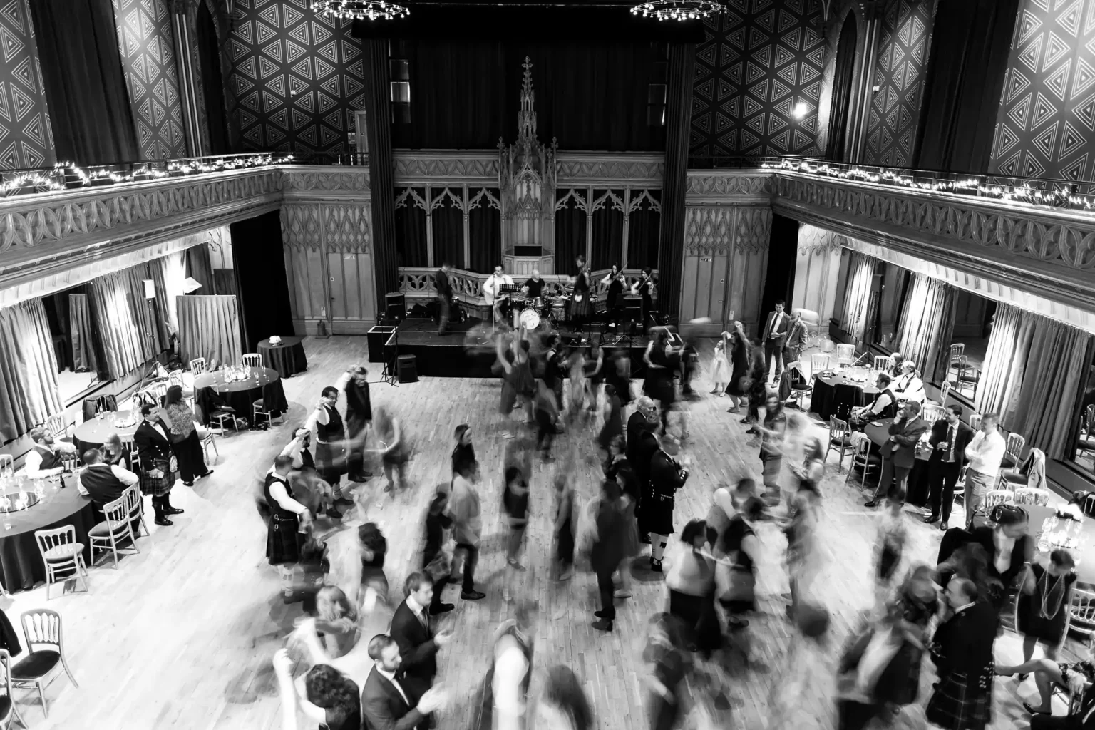 Black and white wedding photography of a ballroom with many people dancing on the floor and musicians on stage. The audience is seated around the room. The image captures motion blur from dancers.