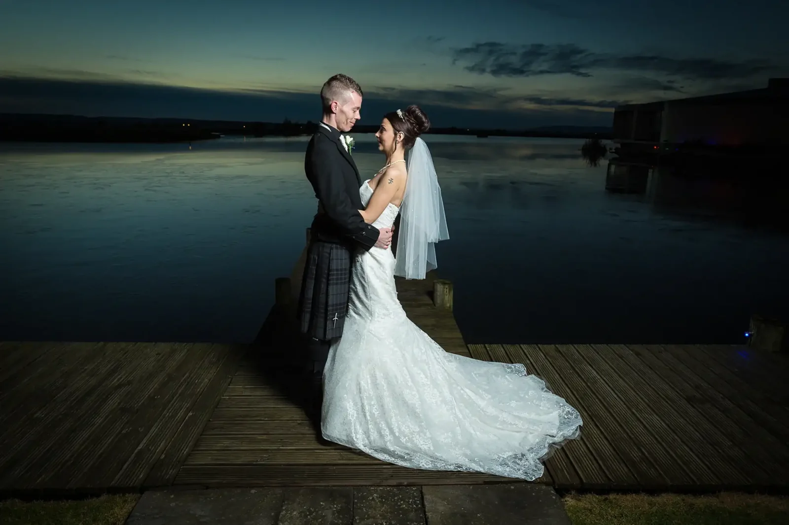 The Vu wedding photographer portrait of a bride and groom on the jetty at Ballencrieff Reservoir at dusk, their figures illuminated against a twilight sky.