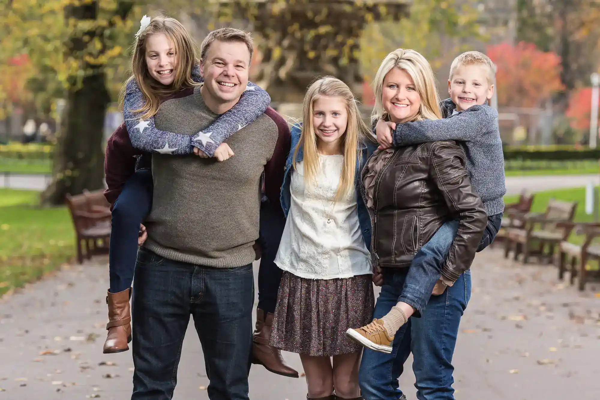A family of four poses outdoors; two children are being carried on the backs of the adults. All are smiling and dressed in casual, autumn clothing. Trees and benches are in the background.