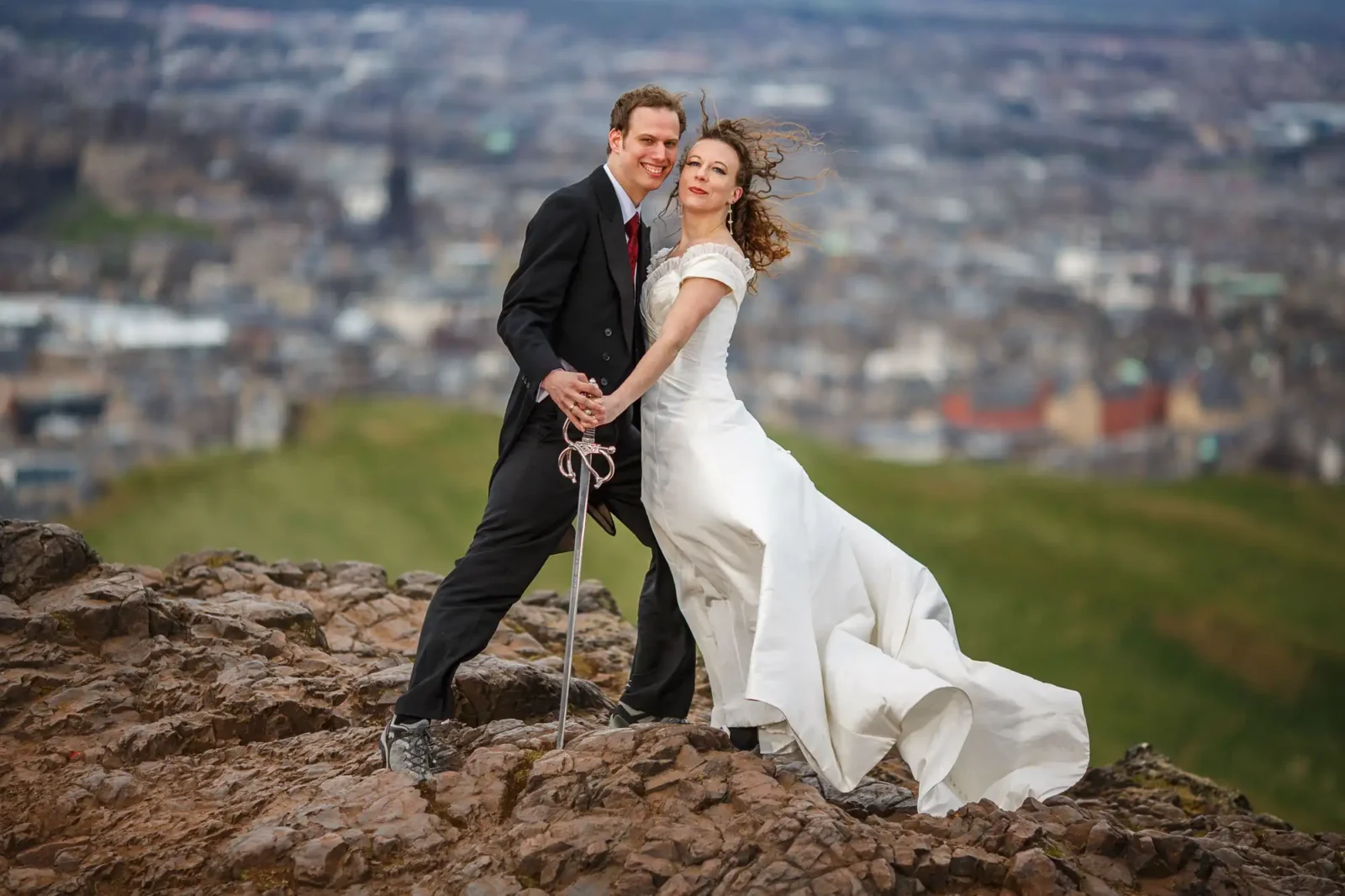 trash the dress photographer image of a bride and groom standing on a rocky hilltop with cityscape in the background, holding hands as the wind blows the bride’s dress and hair.