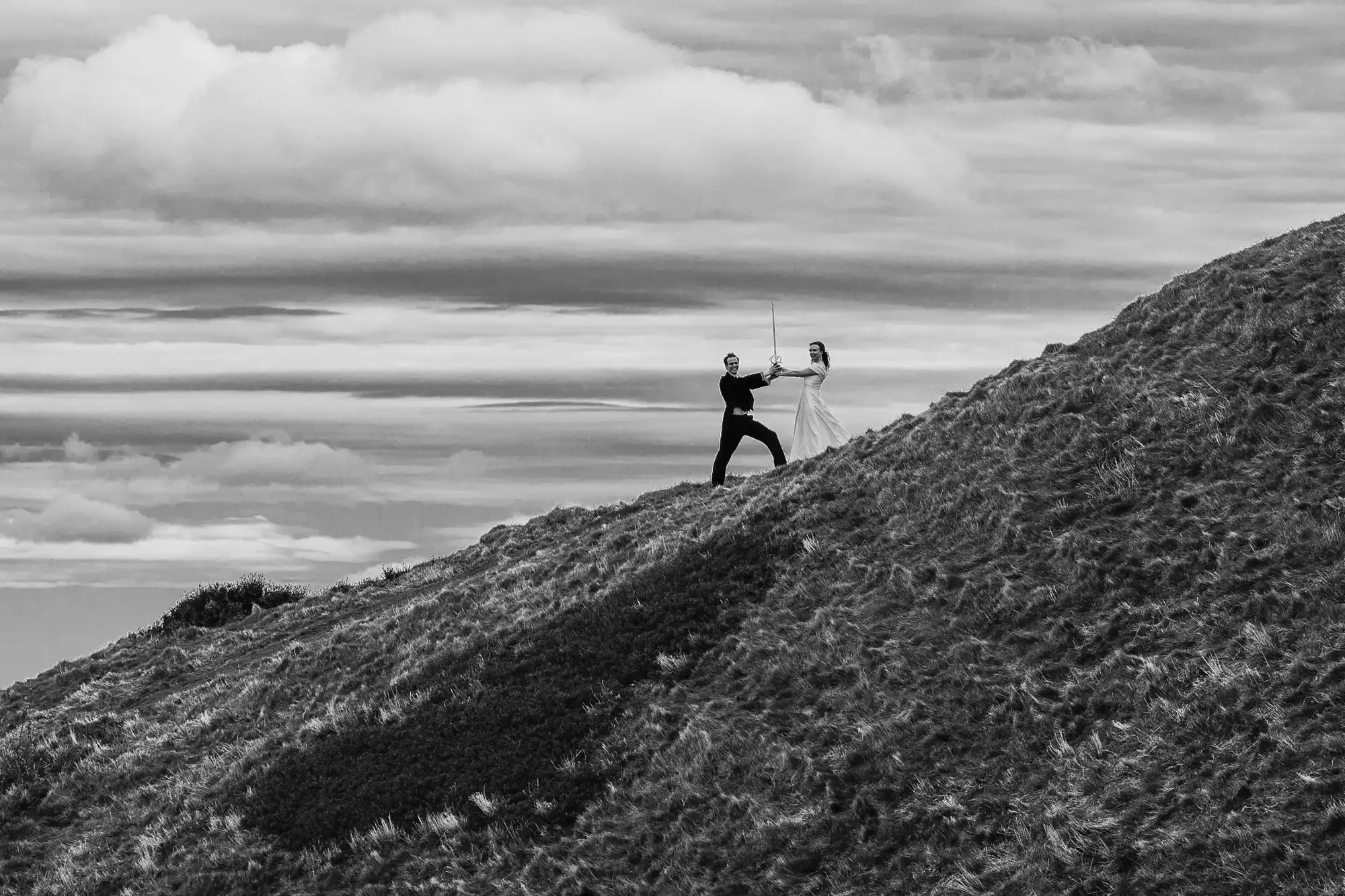 Two people practicing fencing on a grassy hill under a cloudy sky.