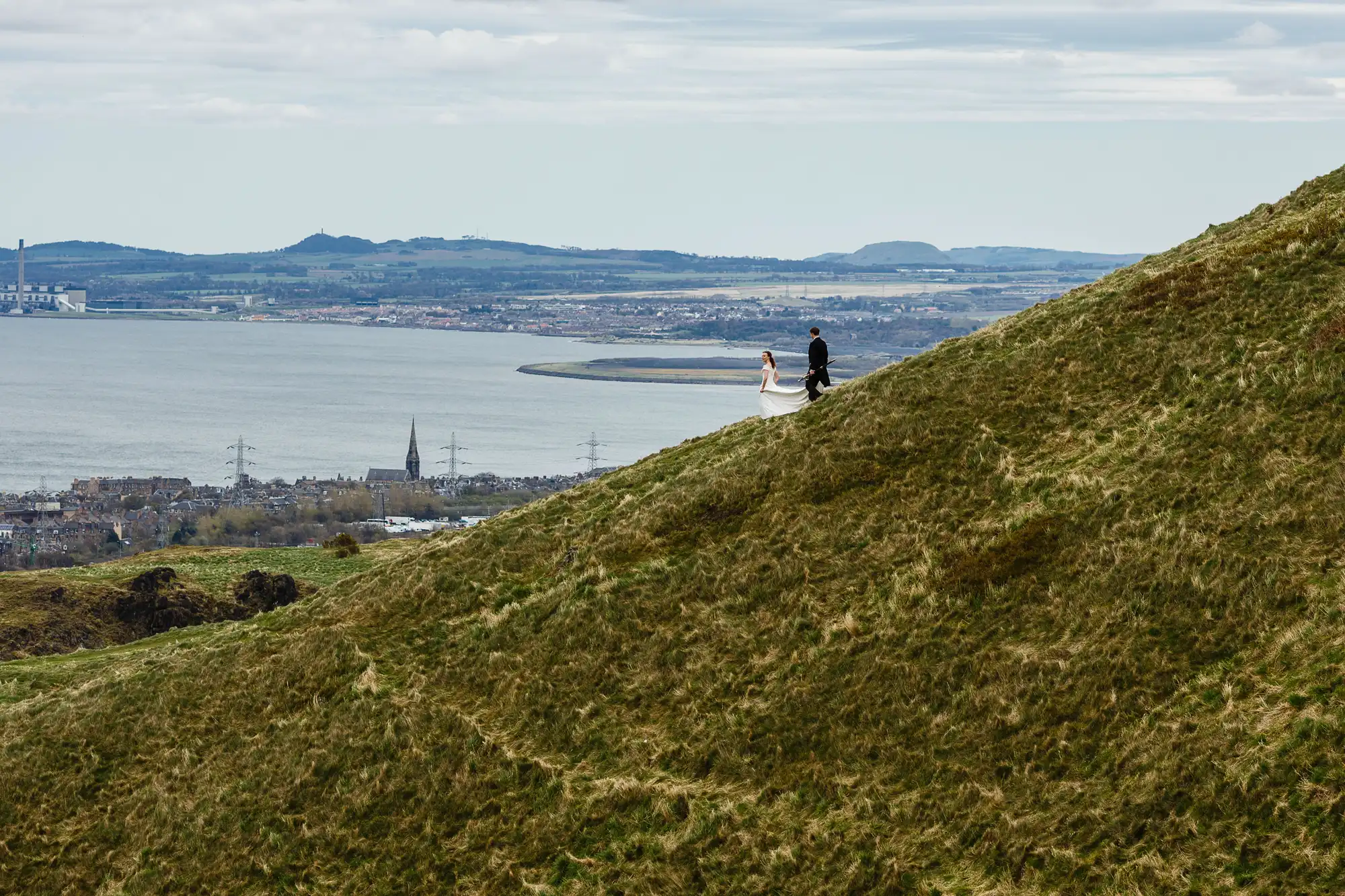 Two people walking on a grassy hill overlooking a city and water in the distance.