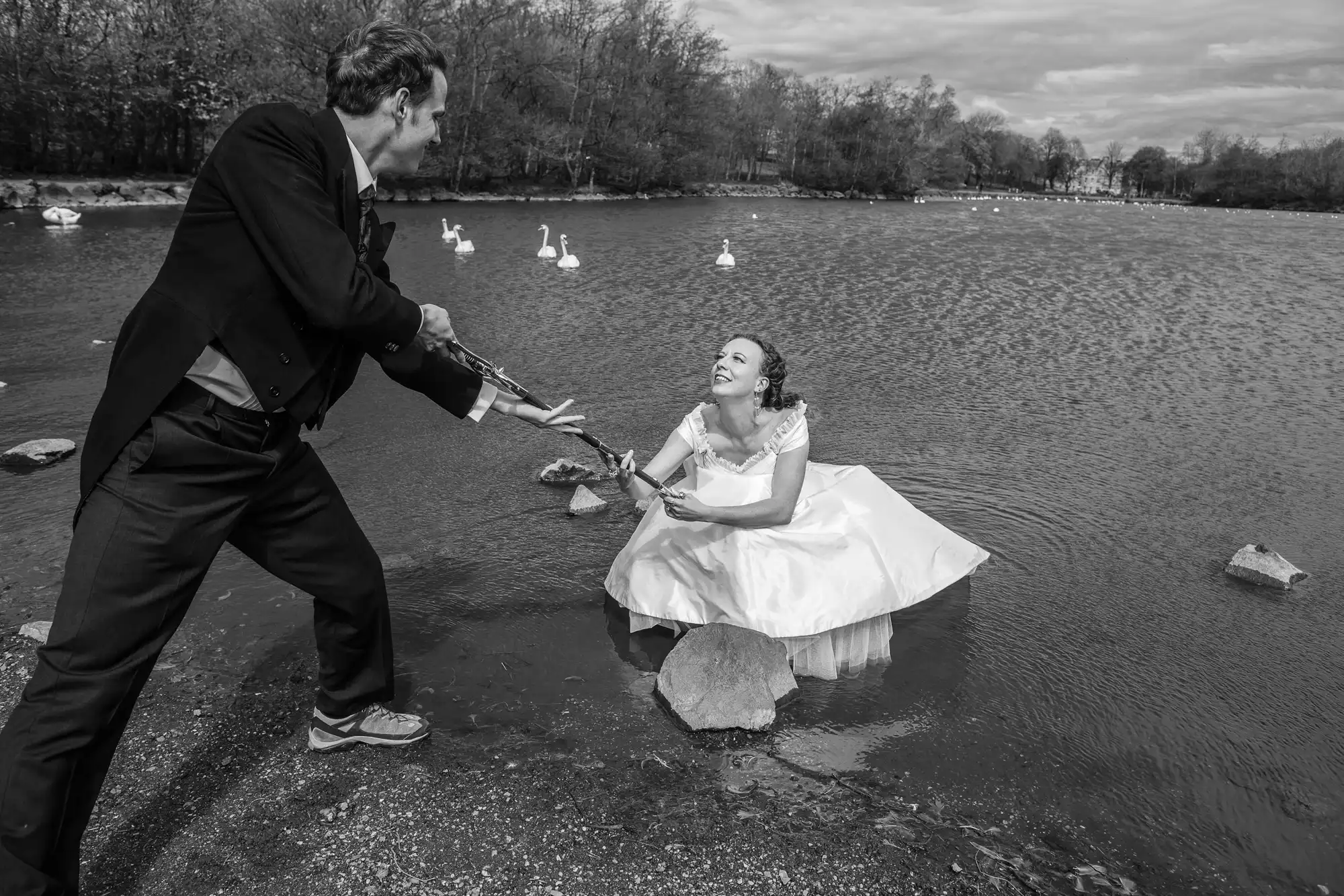 A man in a suit playfully knights a laughing woman in a wedding dress, who is seated on a rock in a shallow lake, with swans in the background.