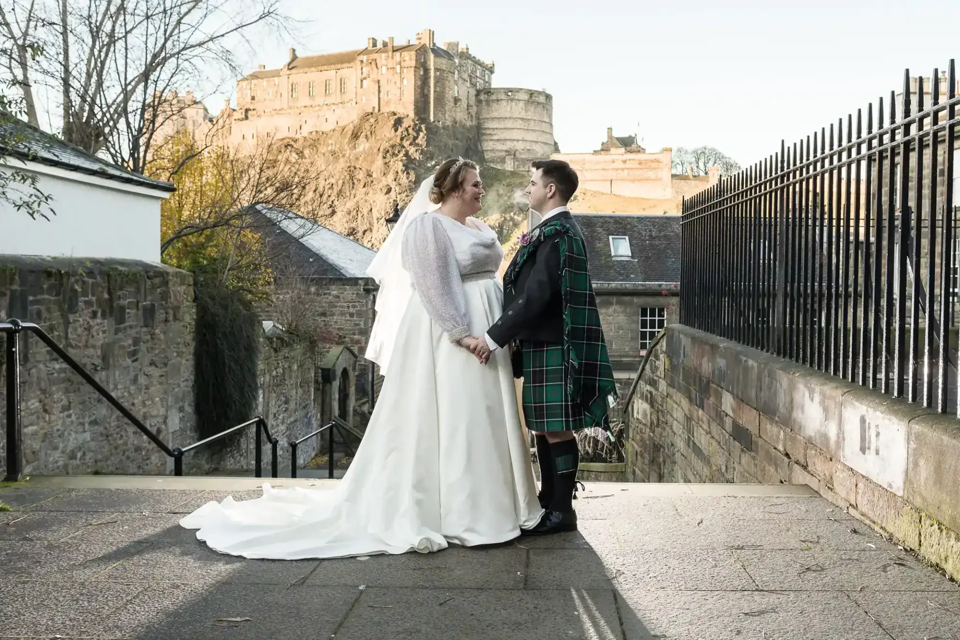 A bride and groom in wedding attire stand facing each other outdoors, with Edinburgh Castle visible in the background.