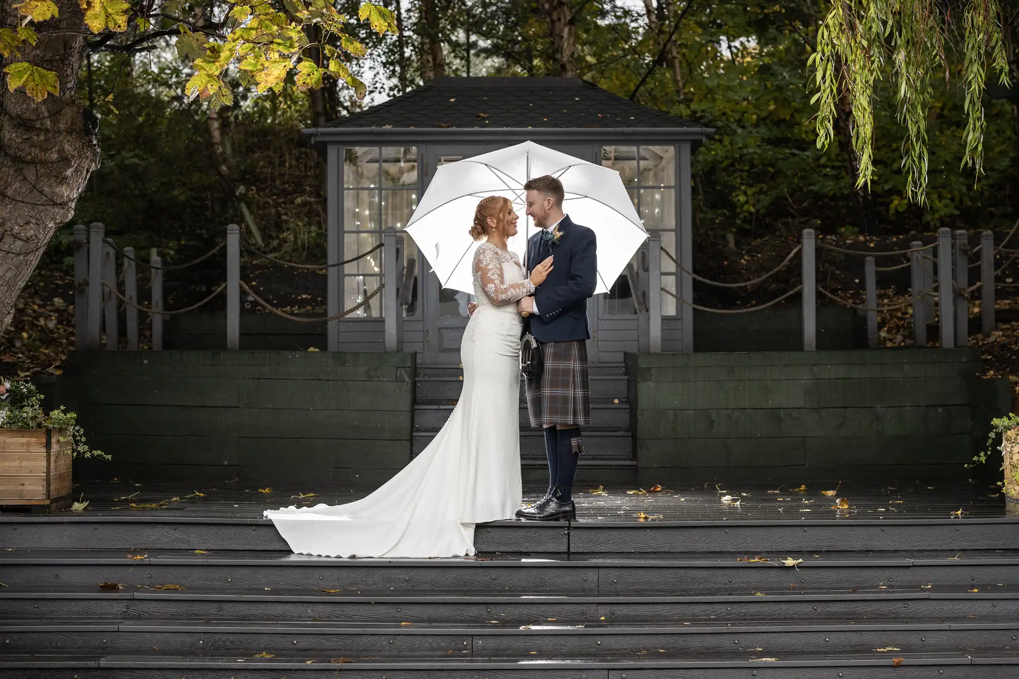 The Venue at Eskmills wedding photographer photo of a bride and groom stand on outdoor steps under a white umbrella, facing each other, with a garden structure and trees in the background.