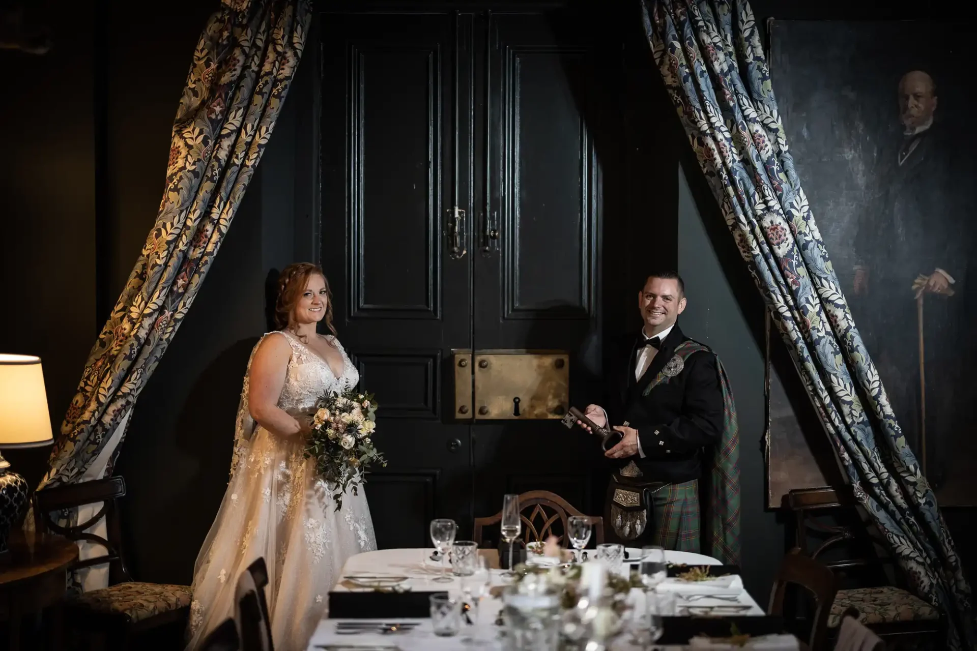 Wardhill Castle wedding photographer: A bride and groom stand smiling in a dimly lit room with floral curtains and a large black door. The bride holds a bouquet; the groom is dressed in formal attire with a kilt.