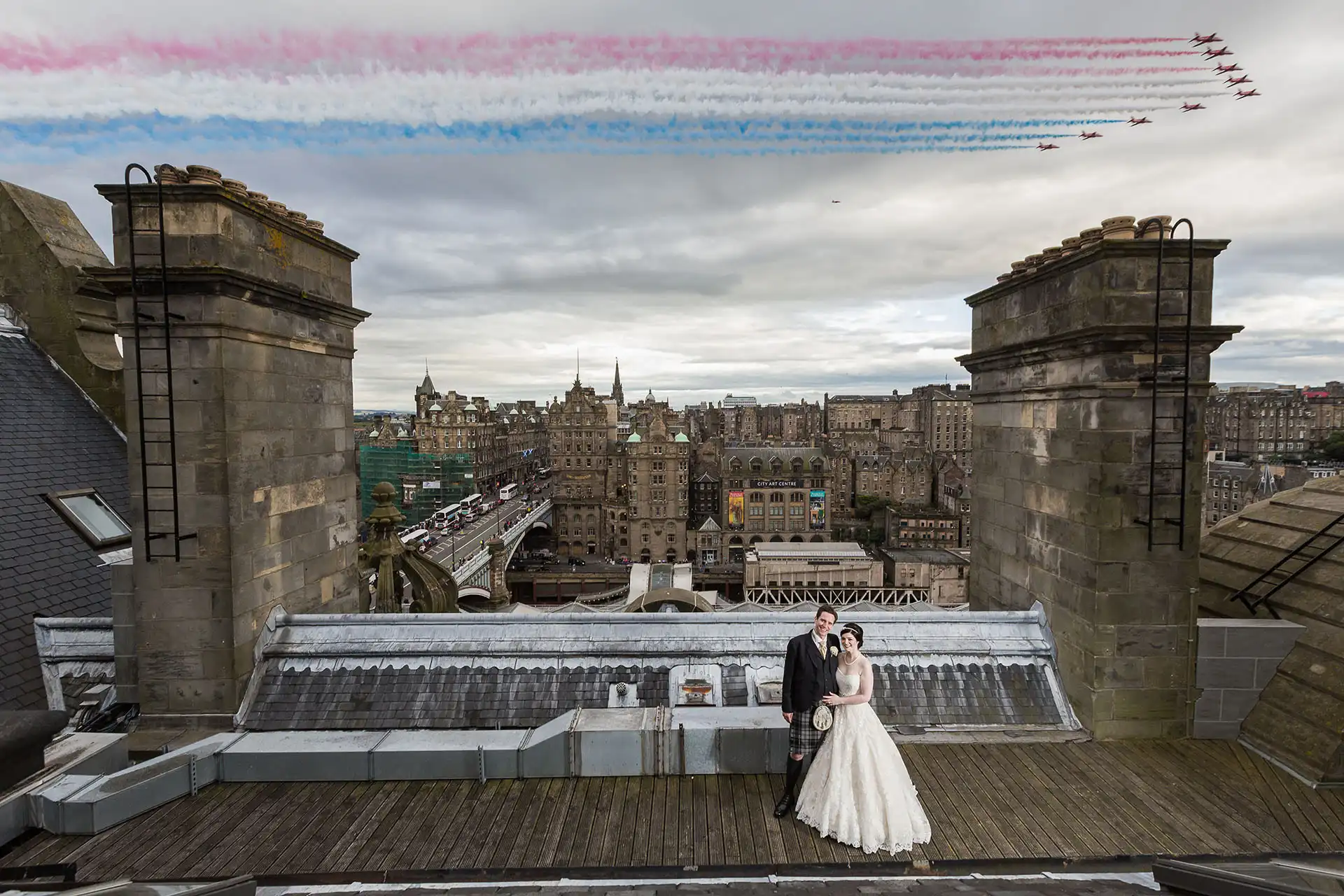 Wedding Photo Of The Year taken on the roof of the Balmoral Hotel: A bride and groom stand on a rooftop overlooking a cityscape, with jets flying in formation and emitting coloured trails in the sky above them.