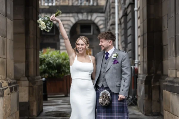 Edinburgh City Chambers wedding photographer image of a bride in a white dress raises her bouquet joyfully while standing next to a groom in a grey jacket and tartan kilt on a stone walkway.