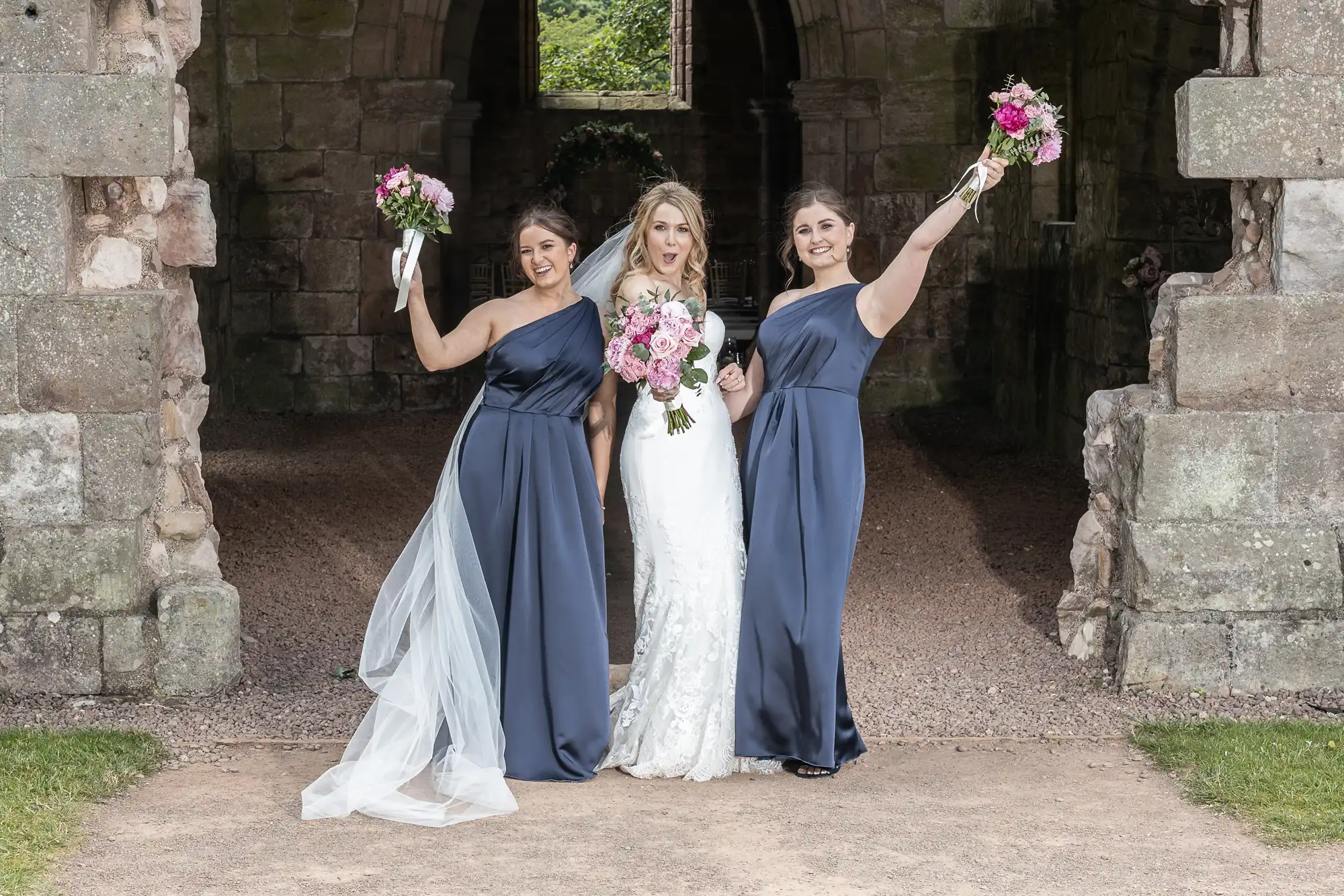 Bride and bridesmaids photography of girls in formal dresses, one in white and two in navy, holding bouquets, pose joyfully at a stone archway.