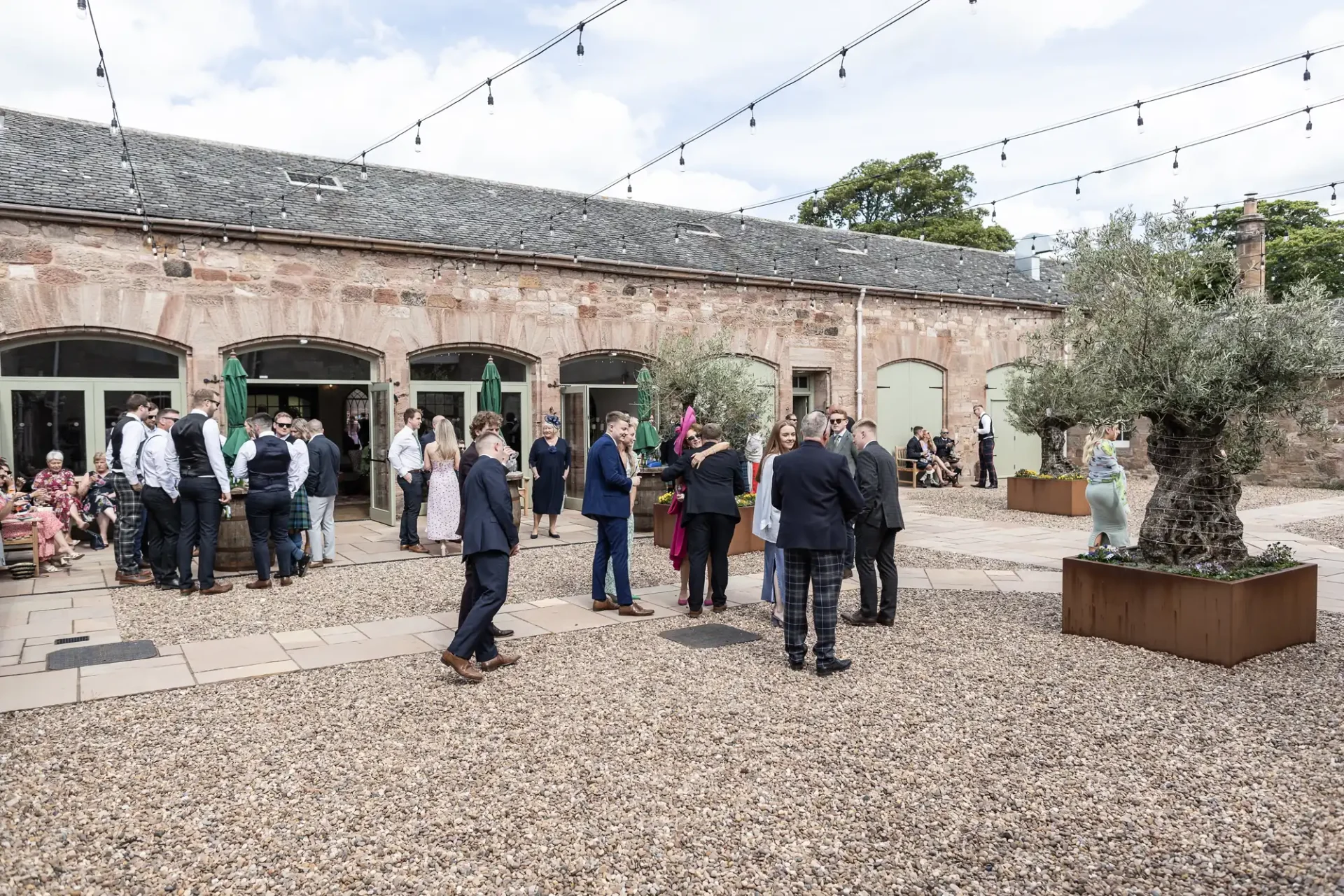 Outdoor social event with adults mingling in Dunglass Stableyard adorned with string lights and trees, some seated under an awning.