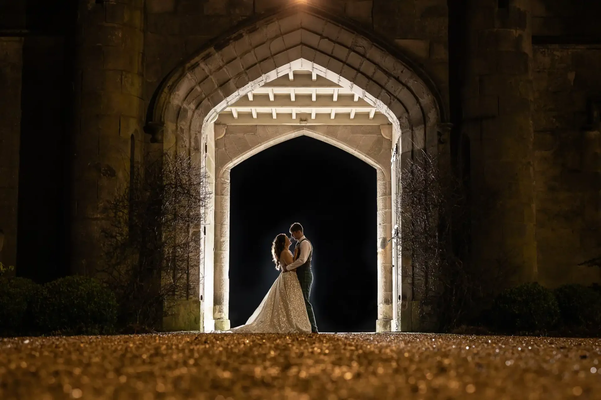 A couple stands embraced under an illuminated Gothic archway at night, with a dark background and a gravel path in the foreground.
