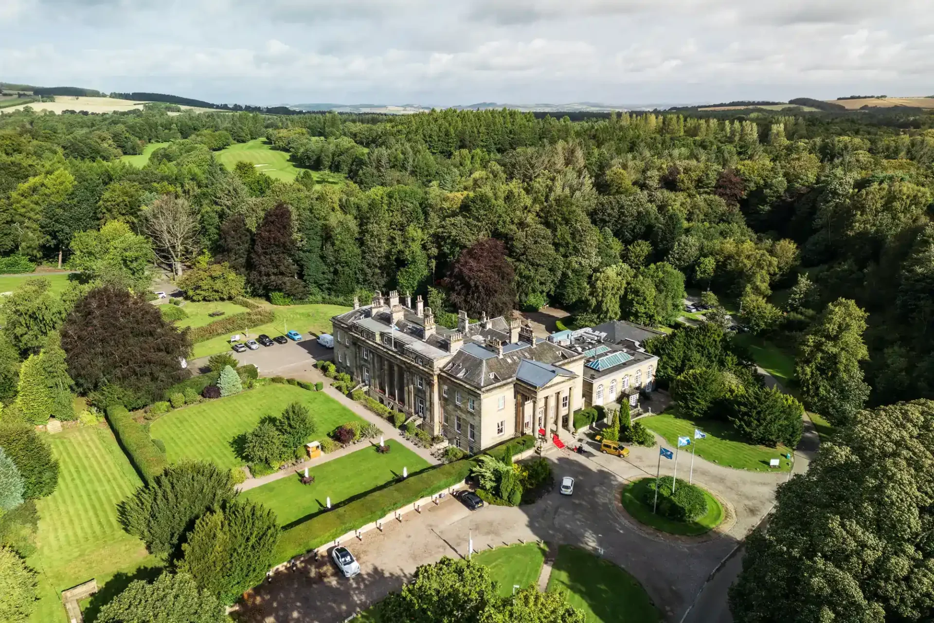 Balbirnie House wedding photographer drone image of the historic mansion with stone walls and many chimneys sits among lush gardens and dense trees. Well-kept lawns, parked cars, flagpoles with blue flags, and winding driveways are visible, with rolling green hills in the distant background.