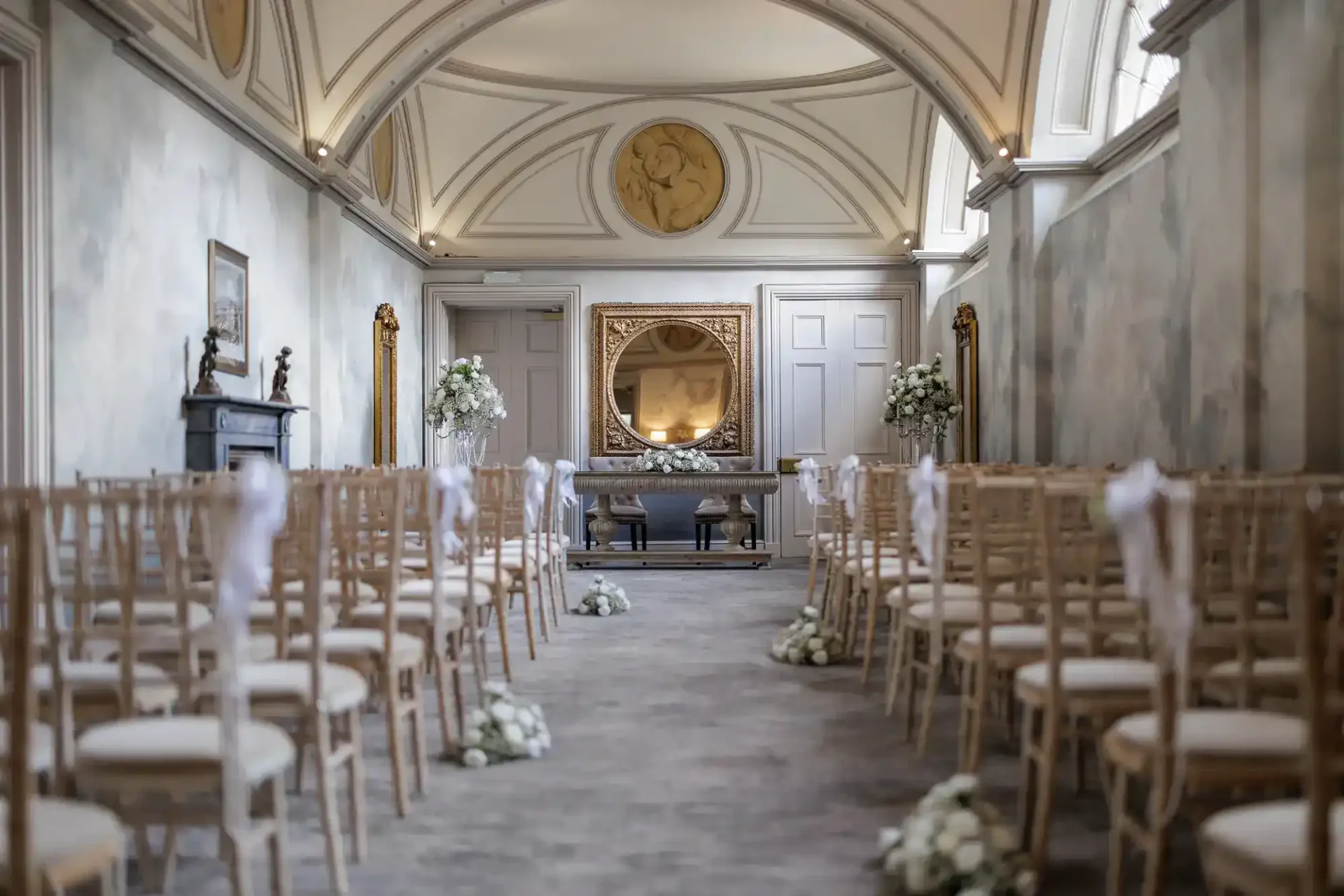 A beautifully decorated, elegant wedding ceremony room, The Long Gallery, with arched ceilings, pale walls, and gold accents. Rows of wooden chairs with white cushions face a table adorned with flowers beneath a large round mirror and classic artwork. White floral arrangements line the aisle.