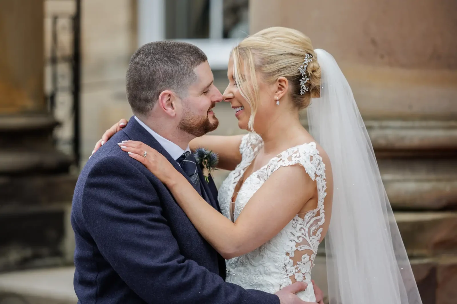A bride and groom, Lisa and Kevin, stand close, smiling at each other and embracing outside Balbirnie House after their wedding. The bride wears a lace dress and veil; the groom wears a suit with a boutonniere.