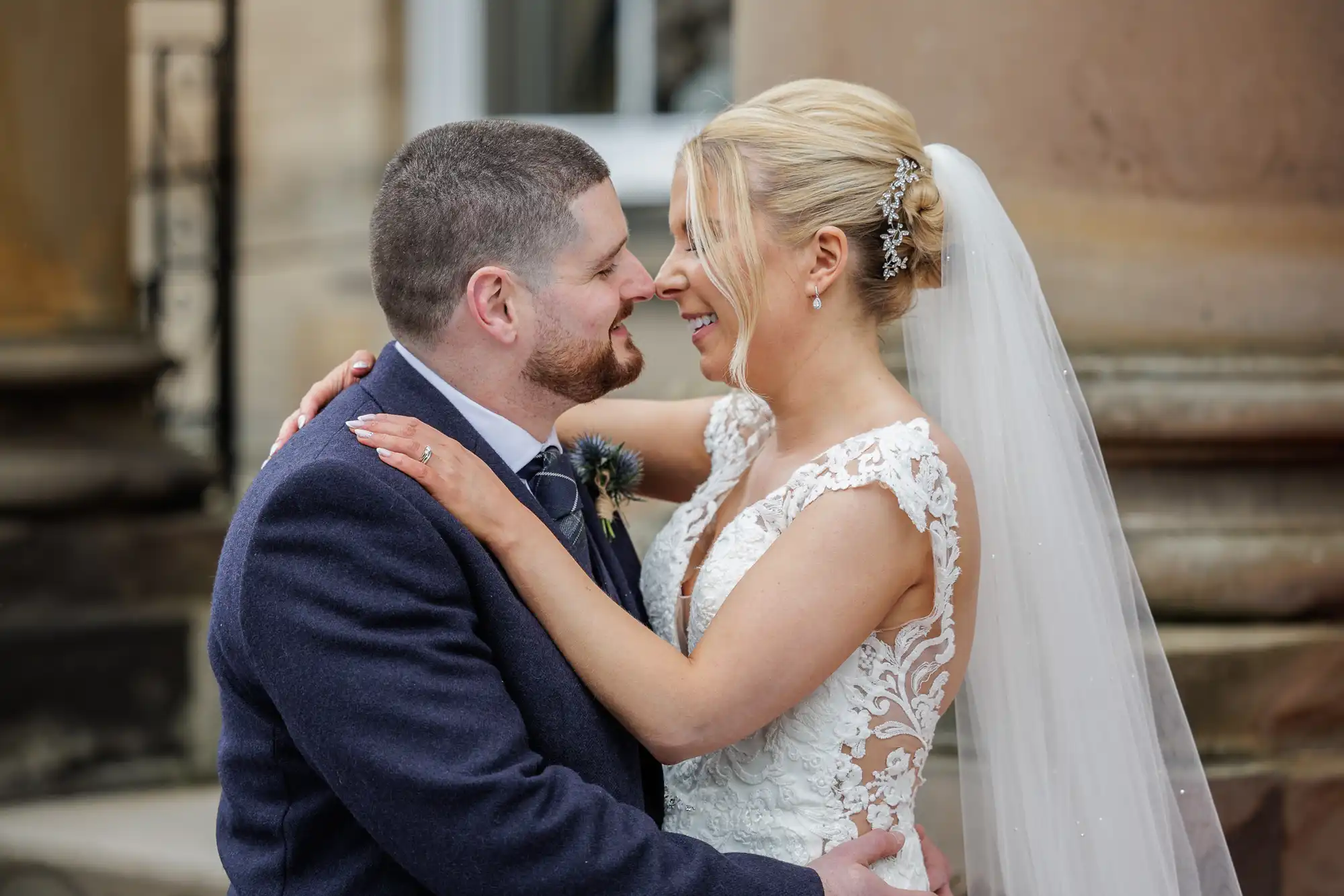 A bride and groom, Lisa and Kevin, stand close, smiling at each other and embracing outside Balbirnie House after their wedding. The bride wears a lace dress and veil; the groom wears a suit with a boutonniere.