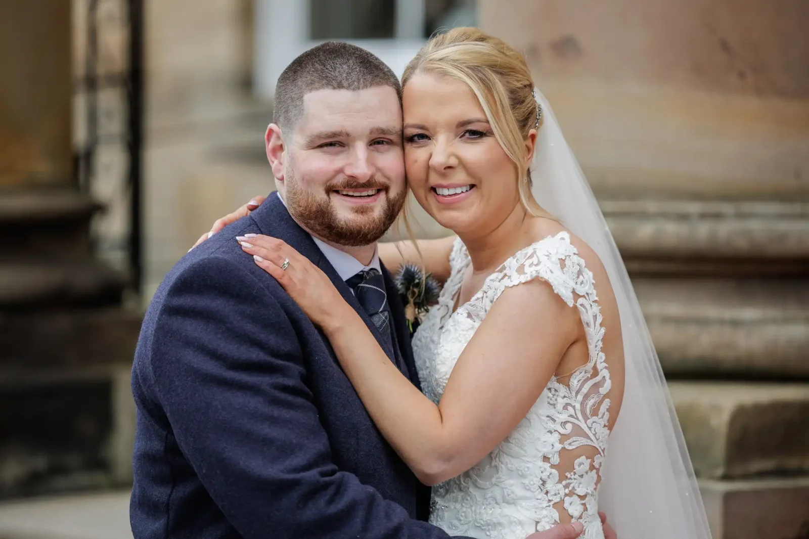 A bride in a white wedding dress and a groom in a dark suit smile and embrace outside a stone building.