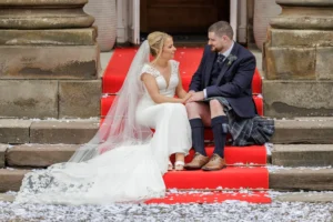 A bride in a white dress and veil sits beside a groom in a kilt on red steps outside a building, surrounded by white confetti.