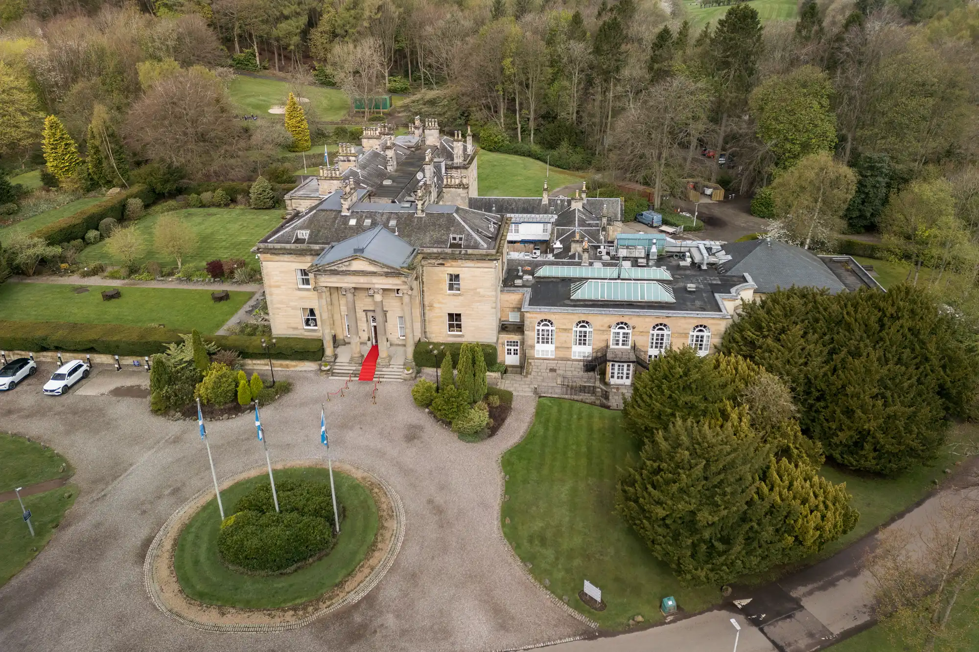 Aerial view of a grand historic mansion with beige stone walls, surrounded by manicured lawns, tall trees, and gardens. Three flagpoles stand in a circular drive at the front. A red carpet leads up to the main entrance with large columns.