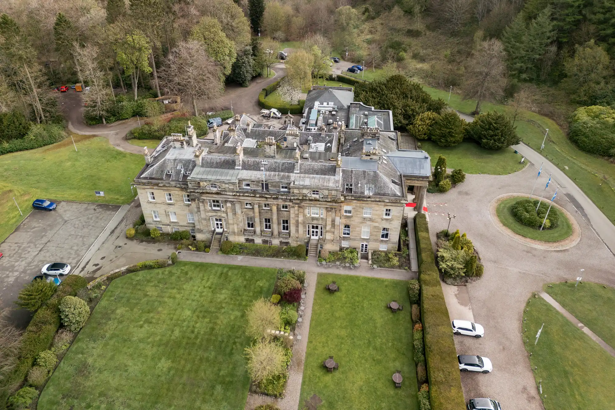 Aerial view of a large historic stone building with a grey roof, surrounded by manicured lawns, pathways, and trees. Several cars are parked near a circular drive to the right. The landscape includes lush greenery and wooded areas in the background.
