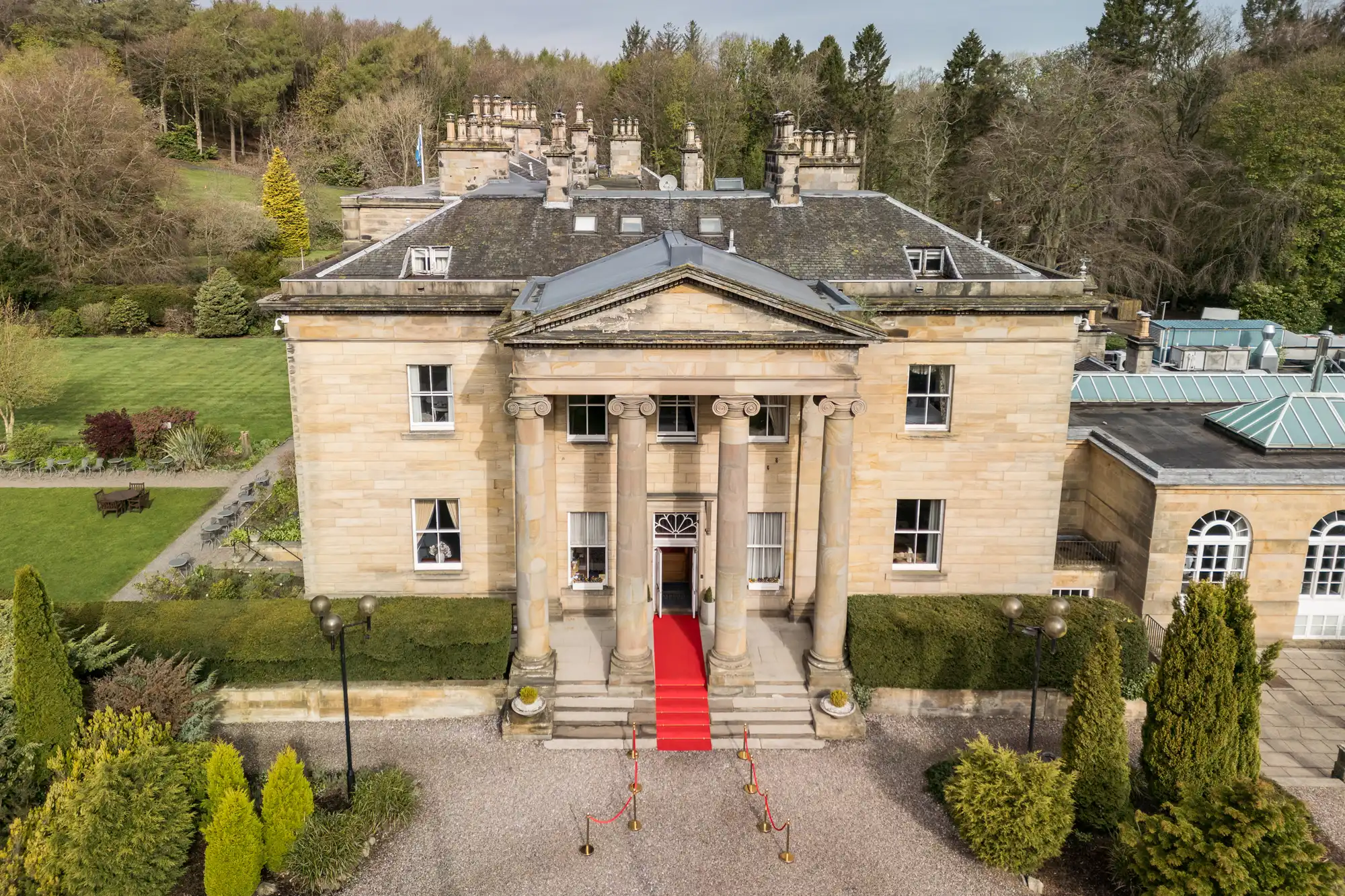 A grand neoclassical mansion with tall columns at the entrance, a red carpet on the steps, ornate railings, and manicured gardens surrounding the building, viewed from above. Green lawns, trees, and shrubs are visible in the background.