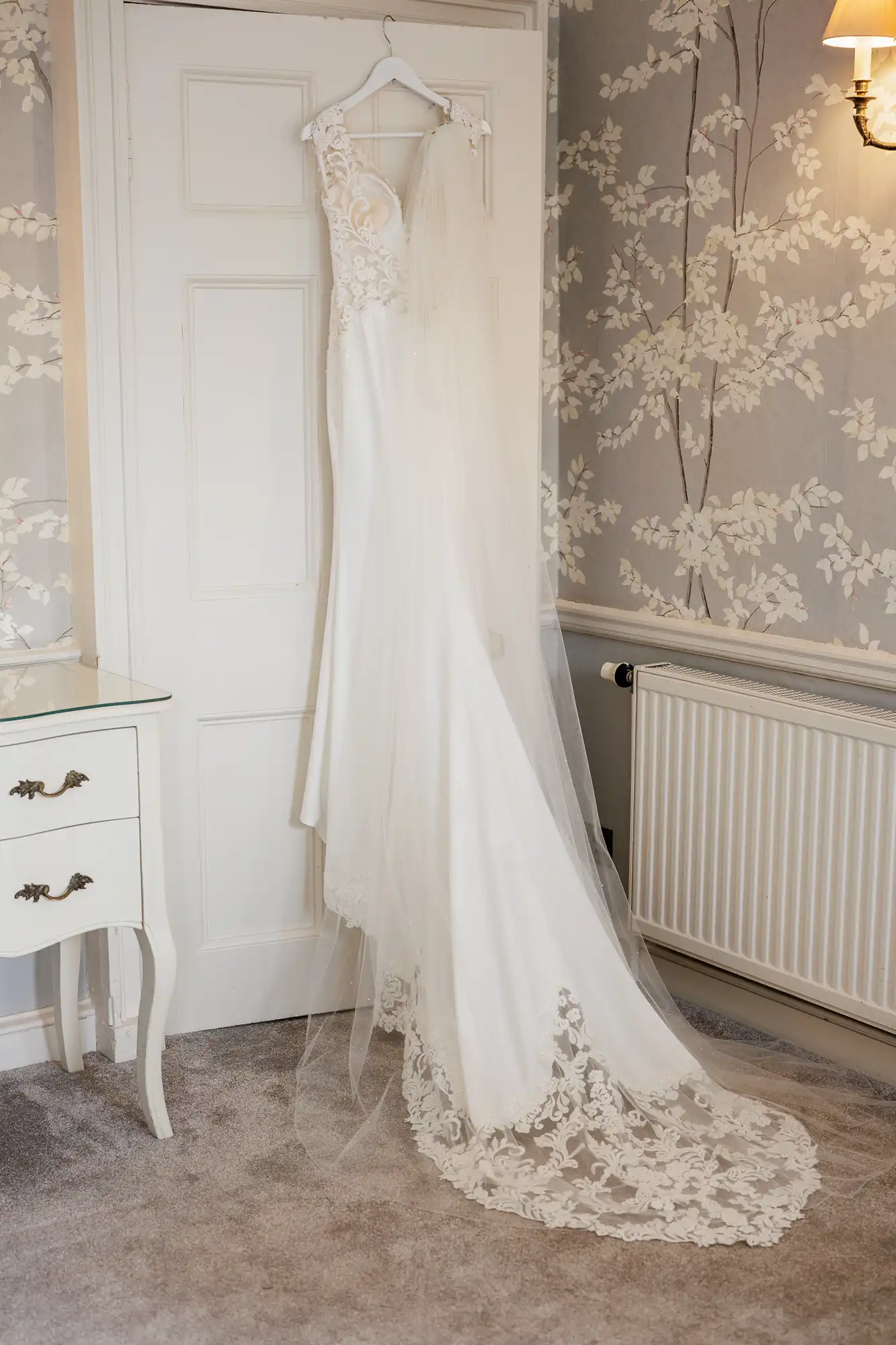 A white lace wedding dress with a long, sheer train and intricate floral details hangs on a door in a softly lit room with floral wallpaper, a radiator, and a small white dressing table with a glass top.