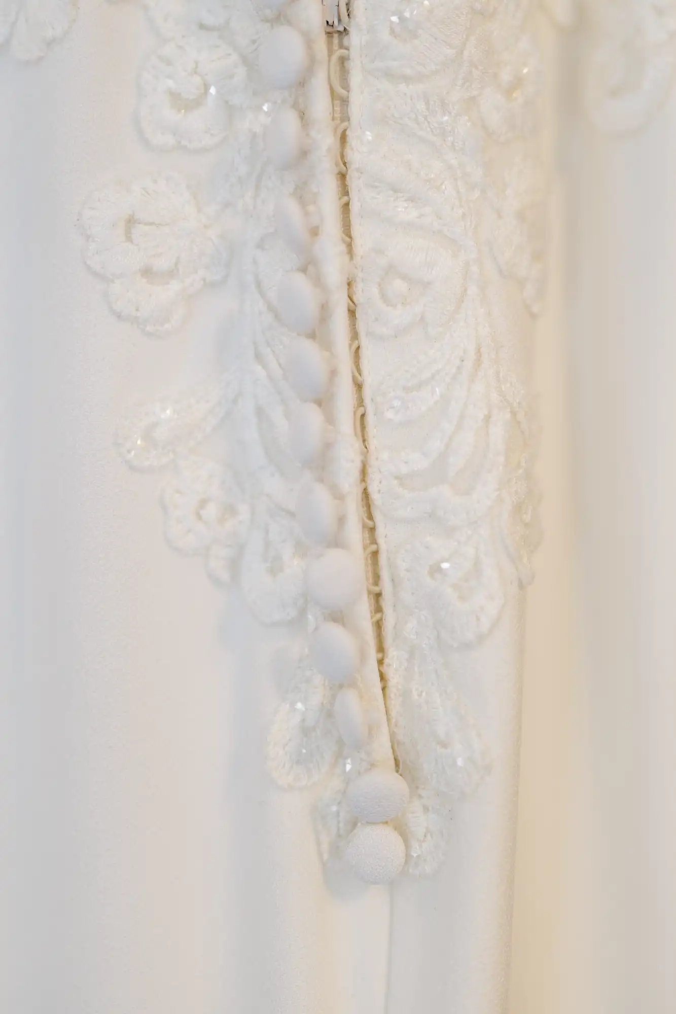 Close-up of a white fabric with delicate floral lace embroidery and a row of covered buttons along the seam, suggesting part of a wedding dress or formal gown. The lace has intricate patterns and a subtle, elegant texture.