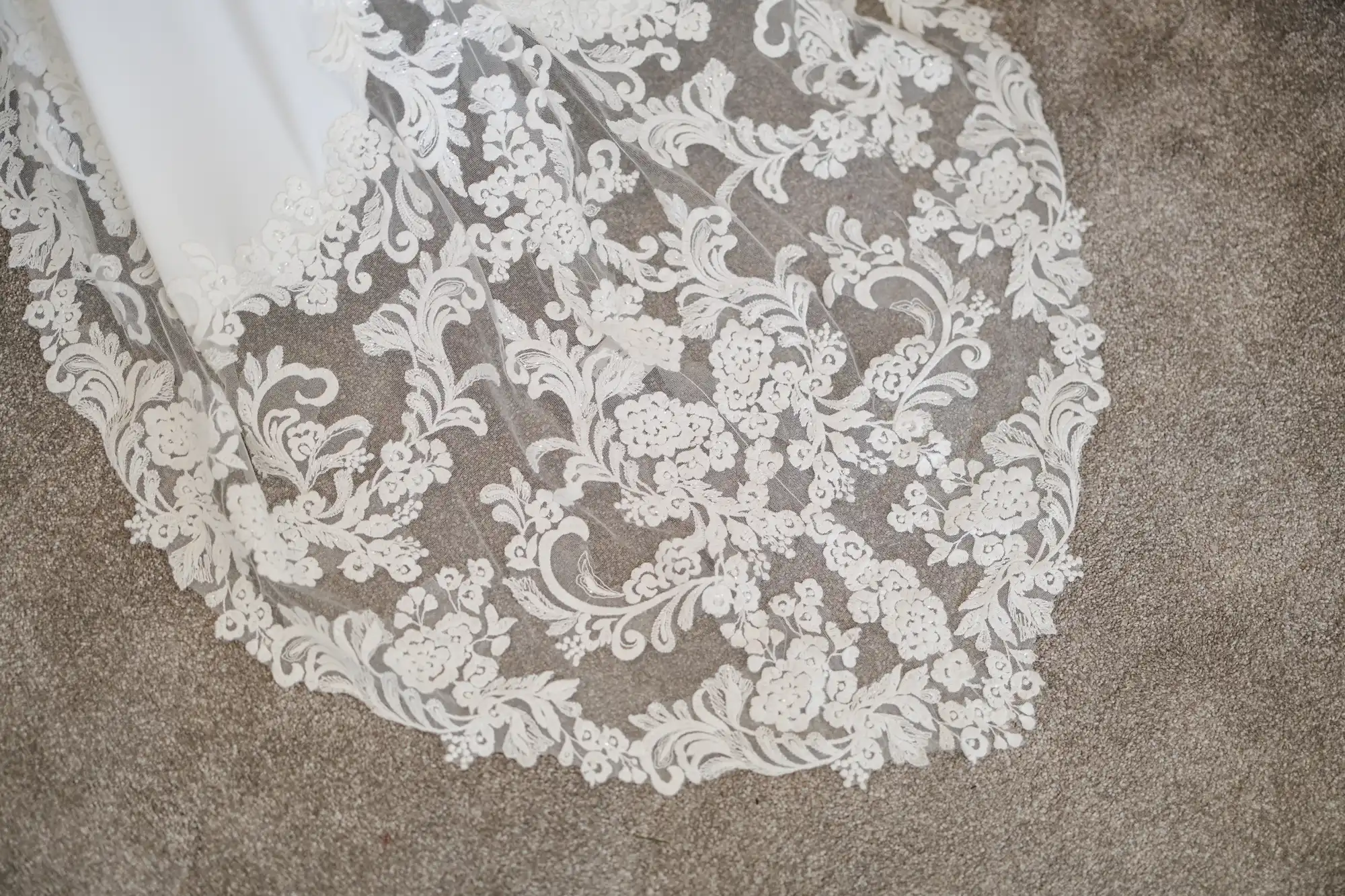 A close-up of a white lace fabric with intricate floral and swirling patterns, likely part of a wedding dress, laid over a light brown textured carpet. The lace has delicate, detailed embroidery and scalloped edges.