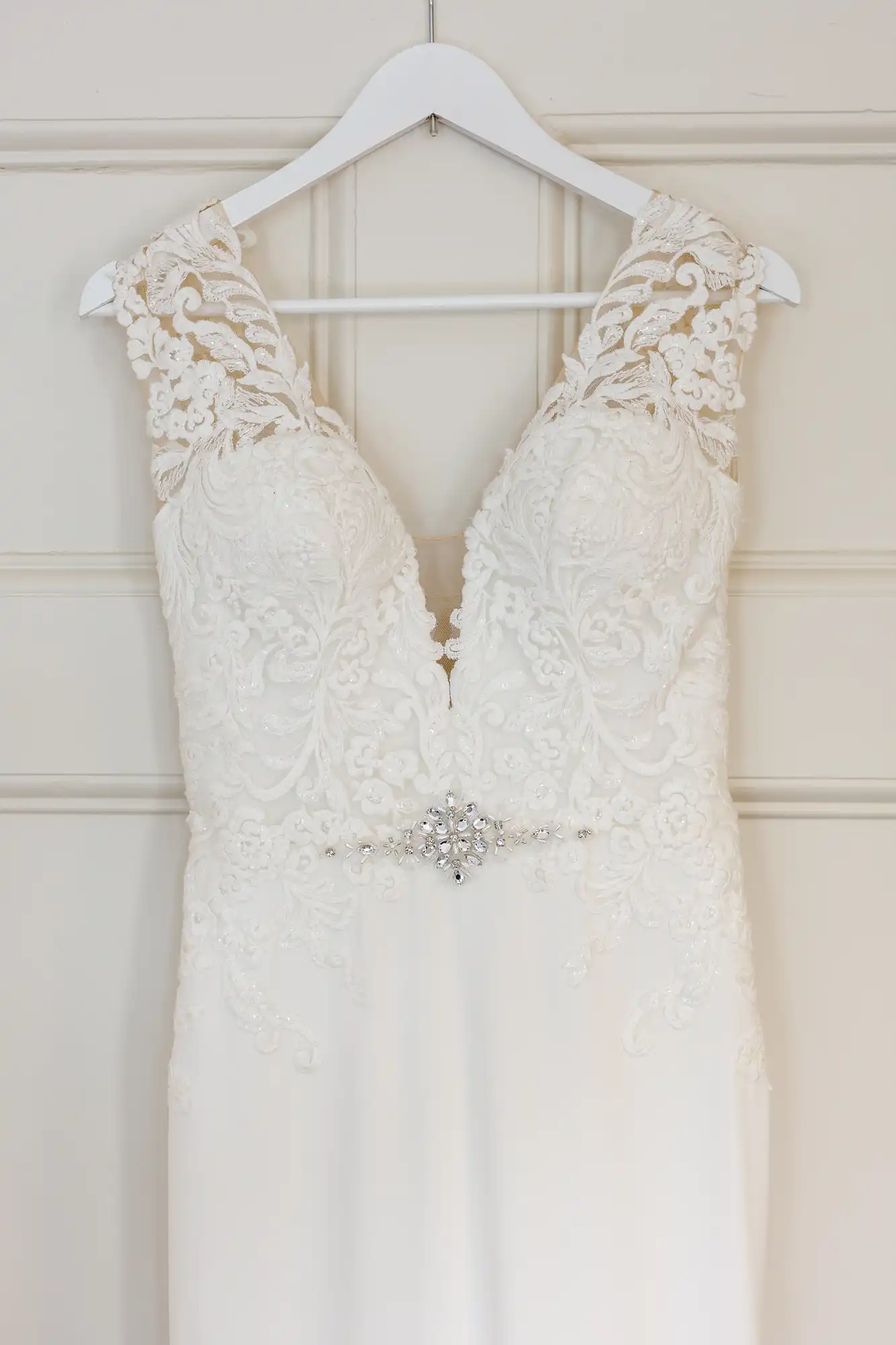 A white lace wedding dress hangs on a white hanger. The dress features a deep V-neckline, intricate lace detailing on the bodice and shoulders, and a jewelled belt accent at the waist. The background is a cream panelled wall.
