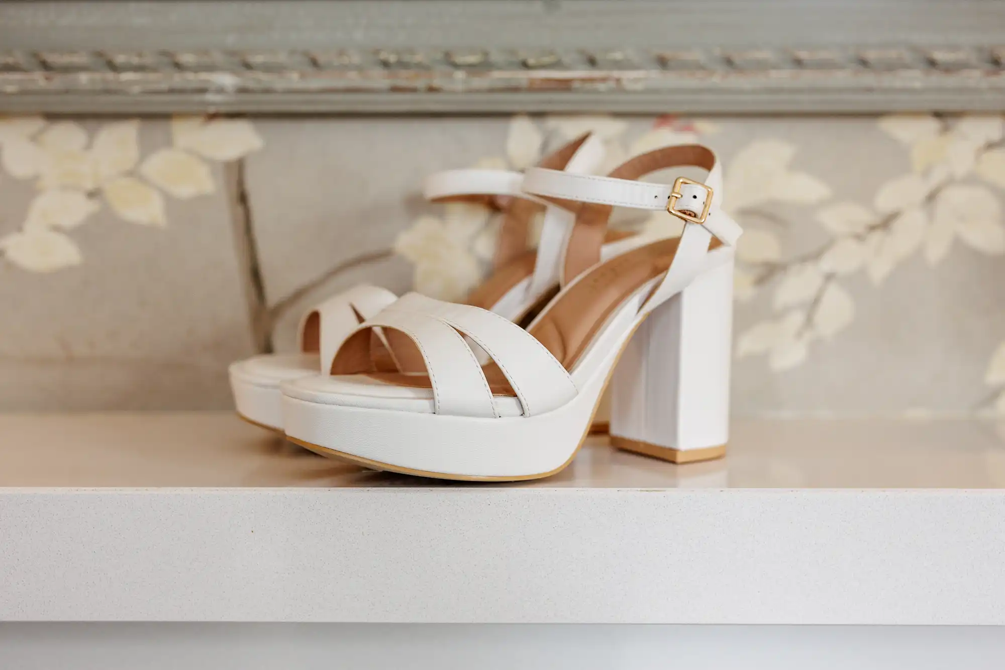 A pair of white high-heeled sandals with thick block heels and ankle straps featuring gold buckles, displayed on a white shelf with a floral-patterned background.