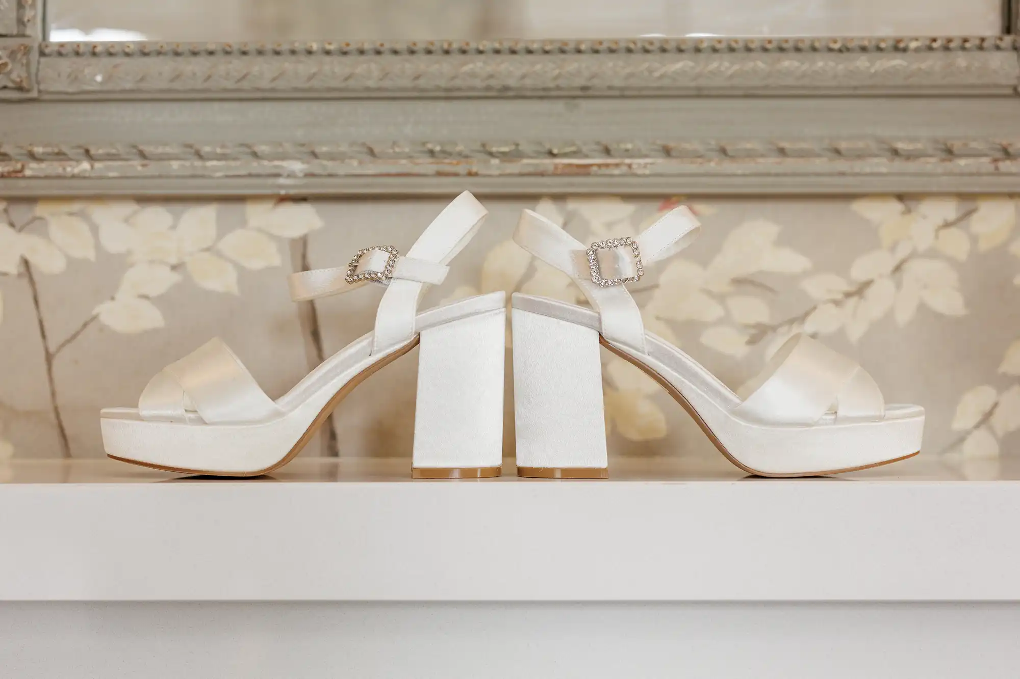 A pair of elegant white satin platform heels with chunky block heels and ankle straps featuring jewelled buckles, displayed back-to-back on a white surface in front of floral wallpaper and an ornate mirror.