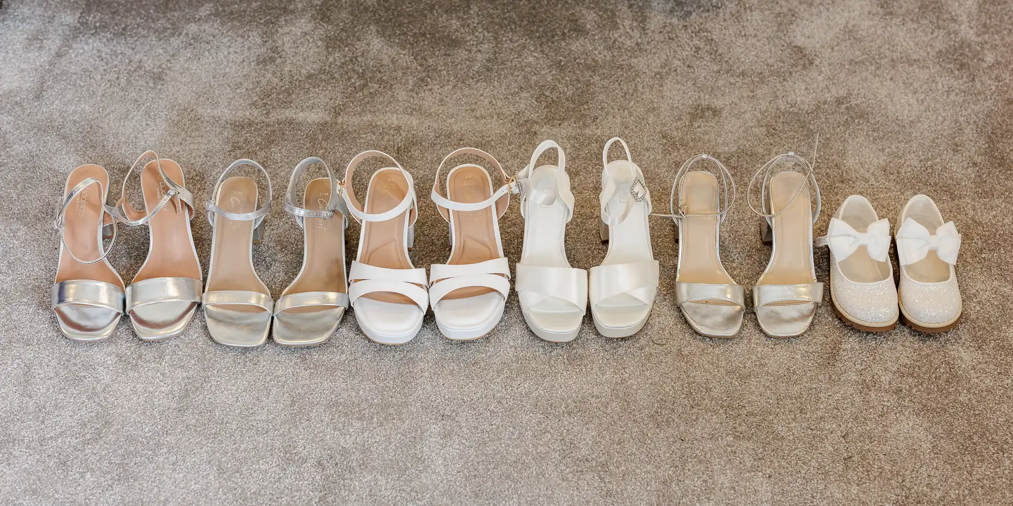 Five pairs of women’s sandals or heels in shades of white and silver are lined up on a carpet. At the end is a small pair of sparkly white children’s shoes with bows, suggesting a family or wedding party.