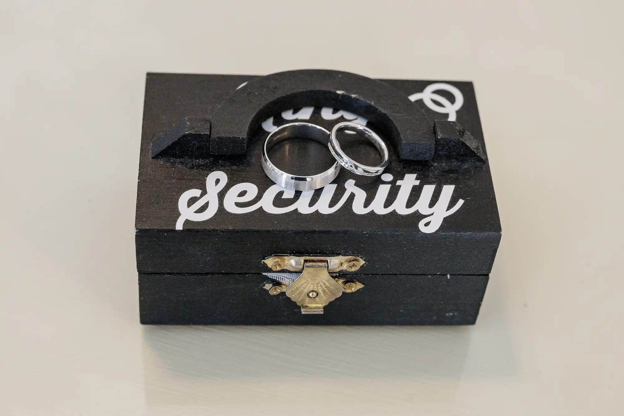A small black box with a brass latch sits on a light surface. The word “Security” is written in white on the lid. Two silver rings rest on top of the box, one plain and one with a black stripe detail.