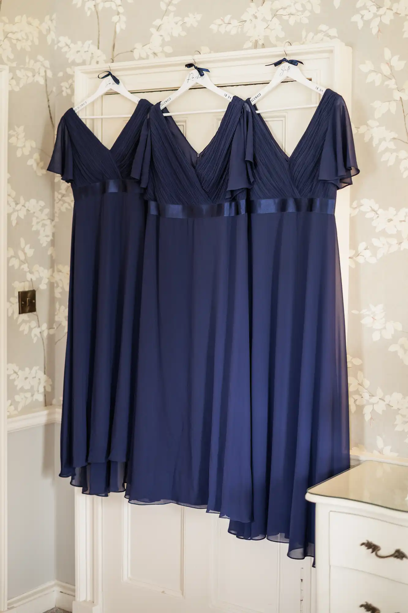 Three matching knee-length, navy blue dresses with flutter sleeves and V-necklines hang side by side on white hangers against light floral wallpaper. The dresses have pleated bodices and flowing skirts, and they are displayed above a white chest of drawers.