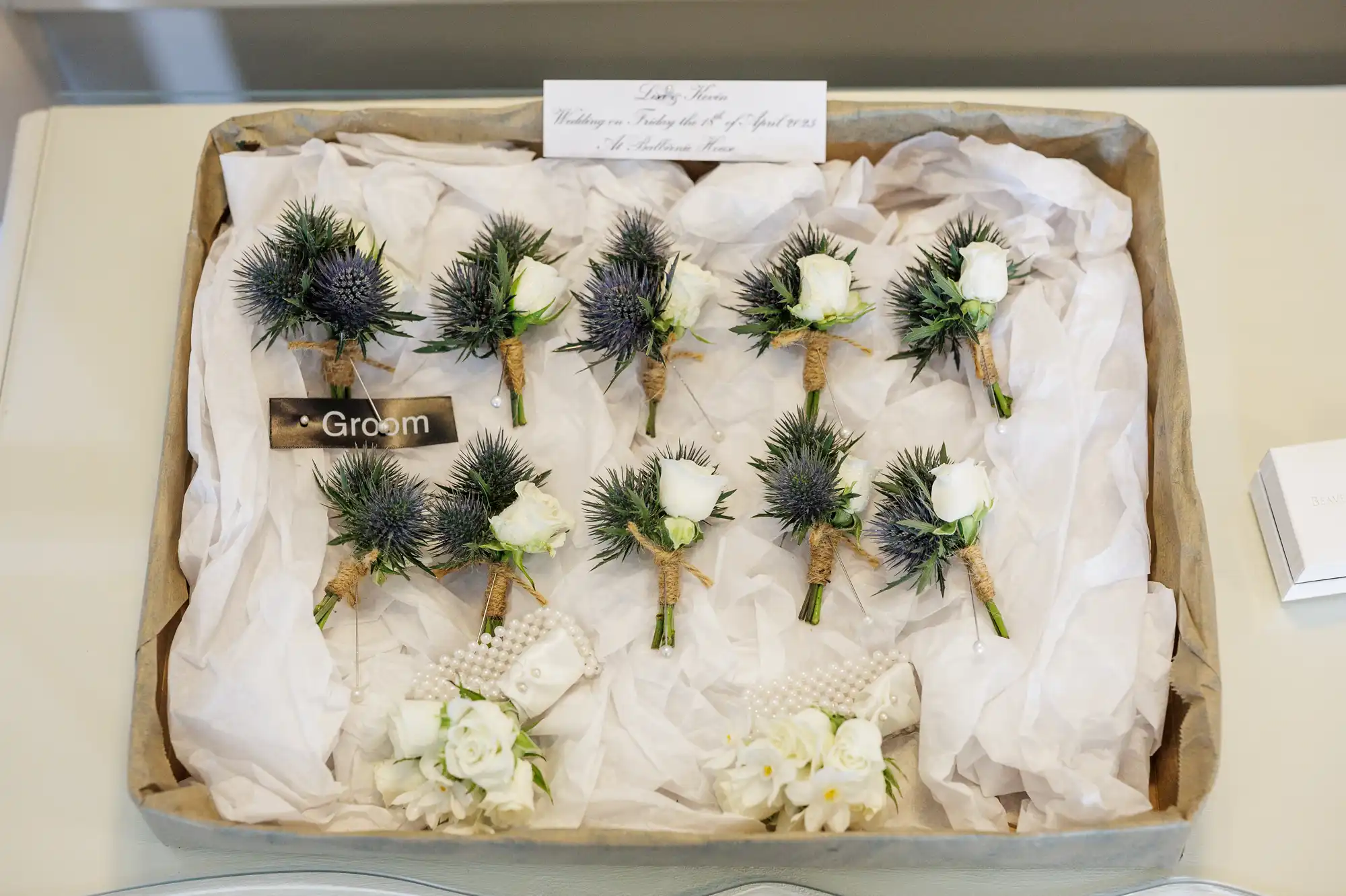 A tray lined with baking parchment holds several buttonholes made of white roses and spiky green thistle. One buttonhole is labelled "Groom." Below the tray are white floral arrangements with pearl accents. A card with handwritten text sits at the top.