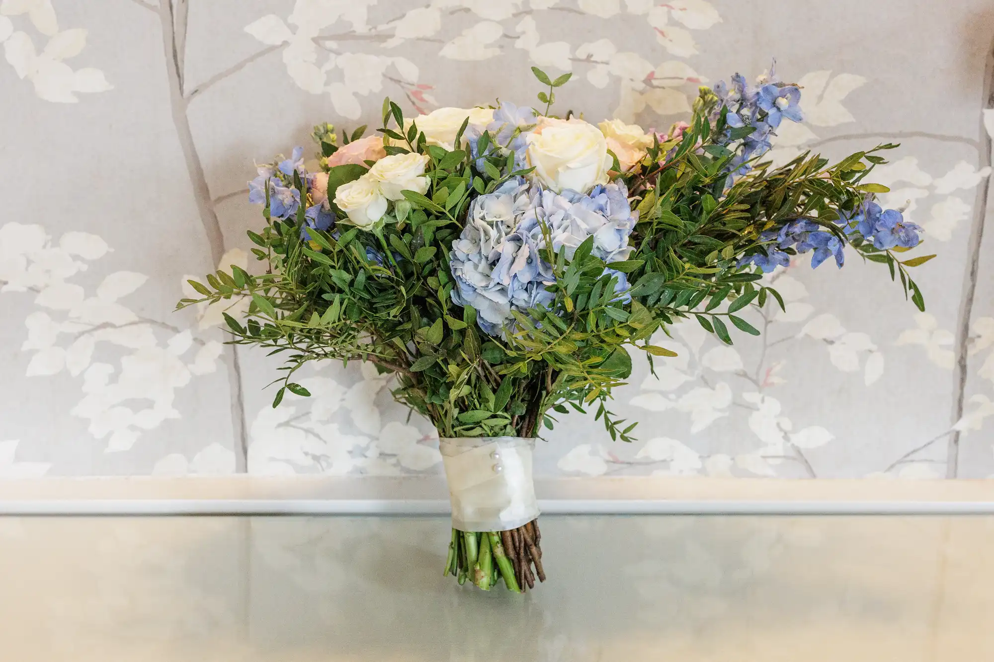A bouquet of white and pale pink roses, blue hydrangeas, and greenery is tied with white ribbon. The bouquet rests on a glass surface, with light grey floral patterned wallpaper in the background.
