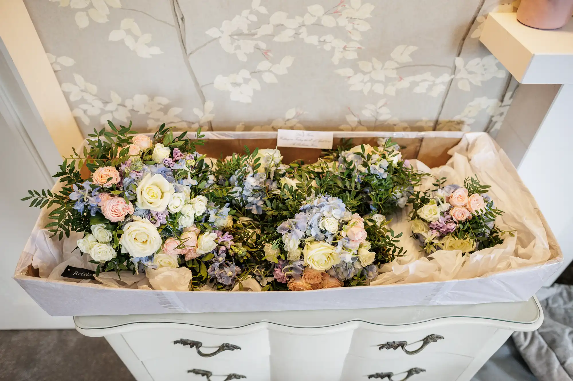 A cardboard box lined with white tissue paper holds several pastel-coloured floral bouquets, including roses, hydrangeas, and greenery, resting on a cream vintage chest of drawers in front of wallpaper with a floral pattern.