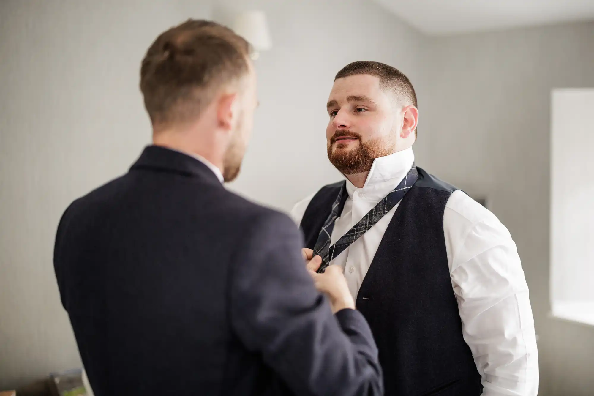 Two men in formal suits stand indoors. One faces the camera, wearing a white shirt and undone tie, while the other, seen from behind, helps him adjust the tie. The room is softly lit with neutral walls and a small window.