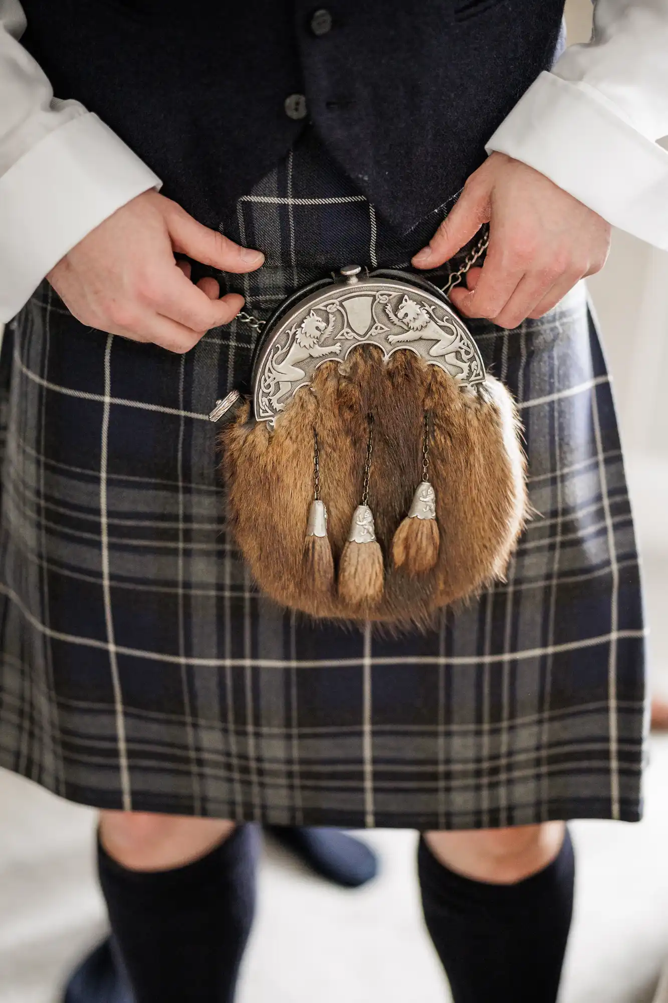 A person wearing a dark tartan kilt and navy waistcoat holds a sporran made of fur with silver embroidery and three decorative tassels. The person's shirt cuffs are visible, and their hands are adjusting the sporran's chain belt.