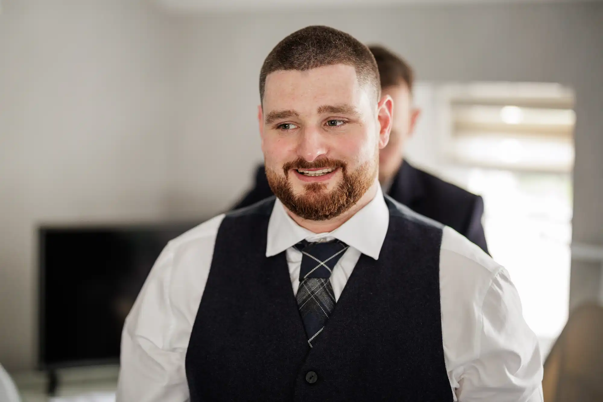 A bearded man with short brown hair smiles whilst wearing a white shirt, checked tie, and dark waistcoat. Another person, mostly out of focus, stands behind him. The room has light-coloured walls, a window, and a TV in the background.